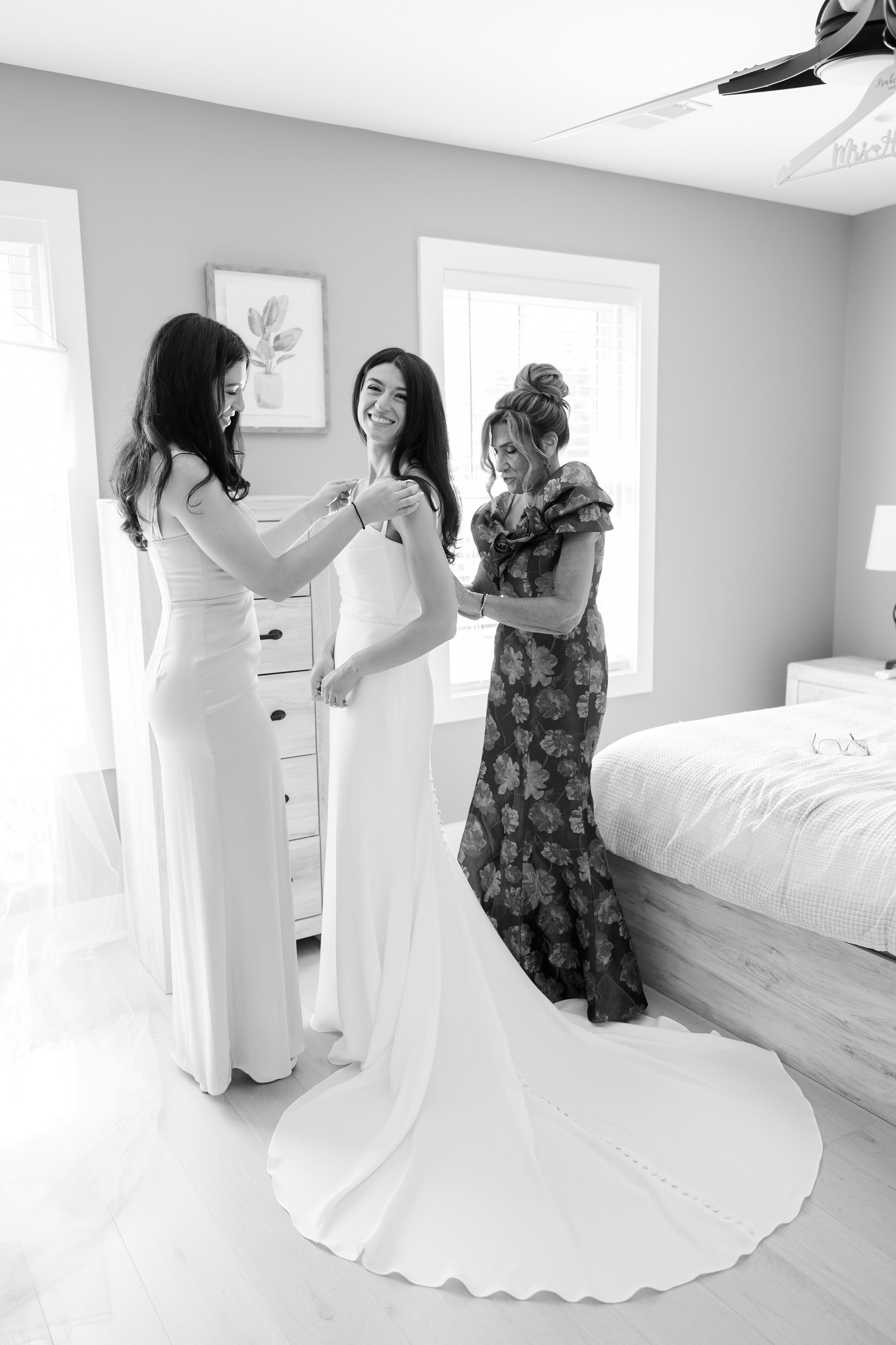 Bride smiles warmly as her sister adjusts her dress strap and her mother carefully fastens the back, a tender moment of love and support as they help her get ready for her wedding day.
