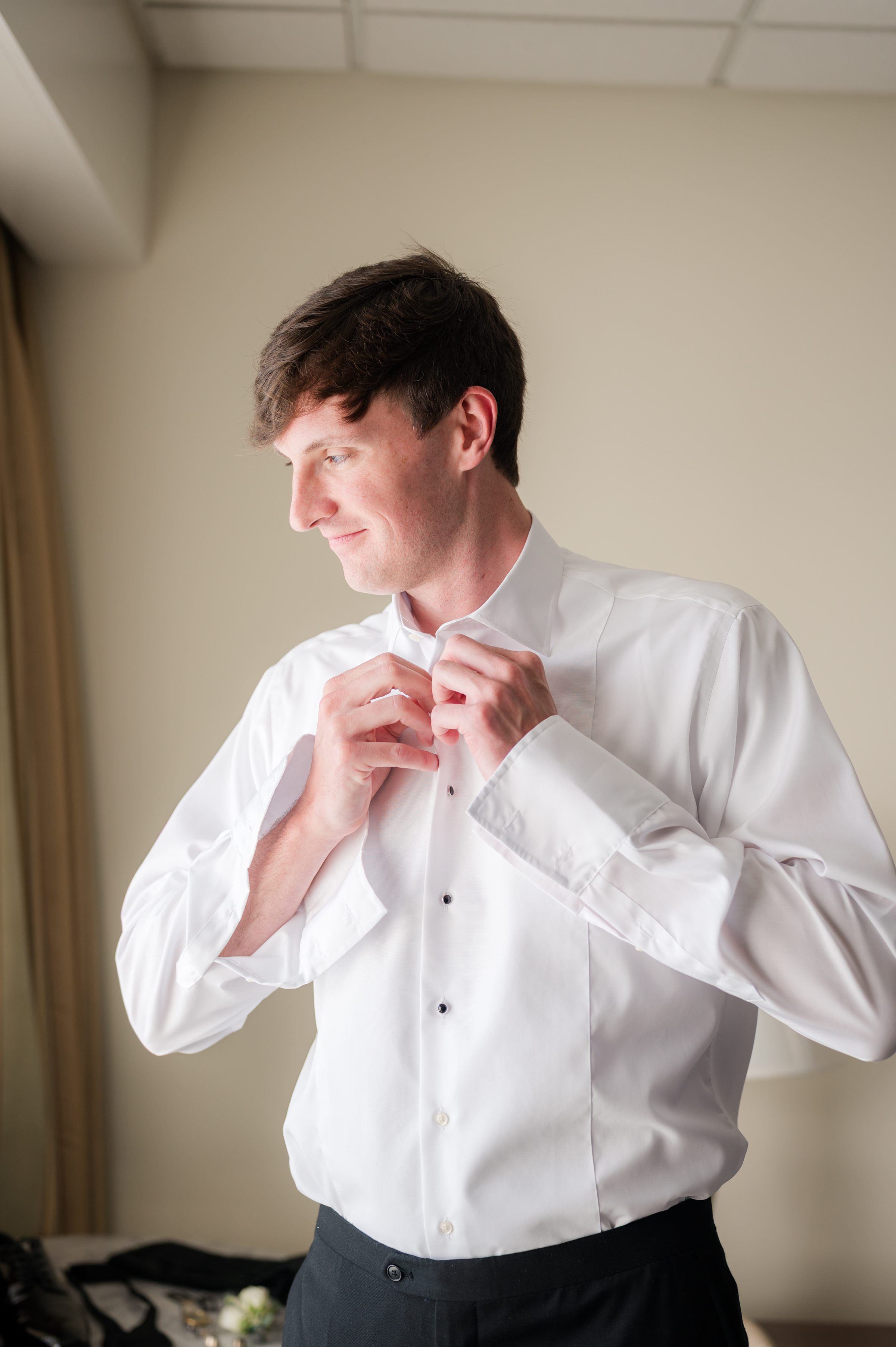 Groom buttons his shirt in soft morning light, preparing himself for the moment he will meet his bride