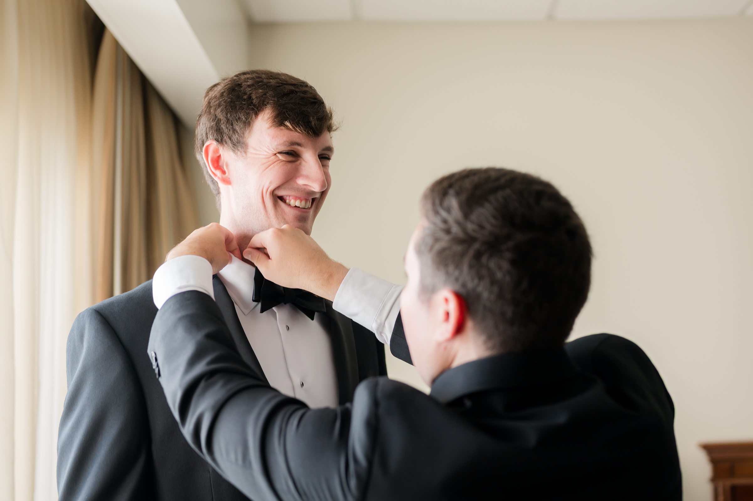 Groom stands still as a groomsman adjusts his bow tie, sharing a lighthearted, supportive moment while getting ready for the wedding.