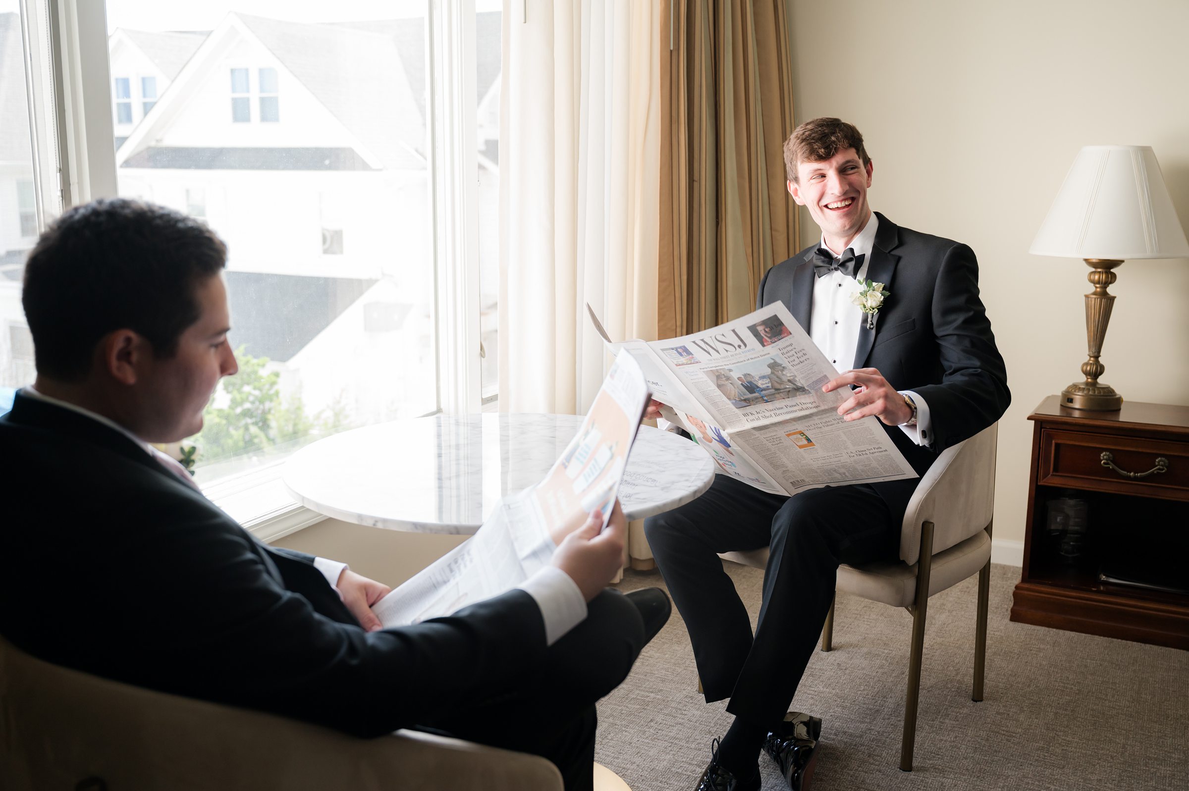 Groom laughs as he reads a newspaper with a groomsman by the window, sharing a relaxed, joyful moment together before the ceremony begins.