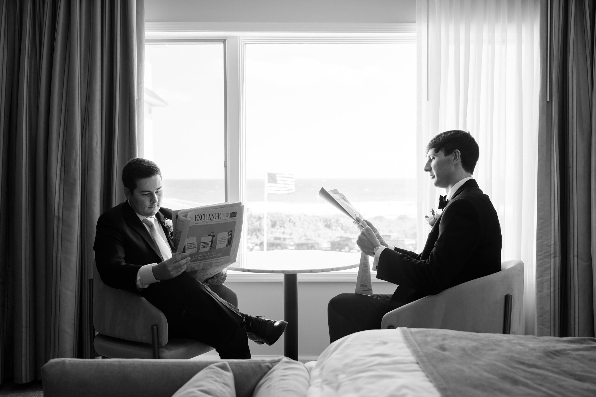 In a quiet, black-and-white moment, the groom and his groomsman sit by a sunlit window reading newspapers, sharing a calm pause before the wedding day unfolds.