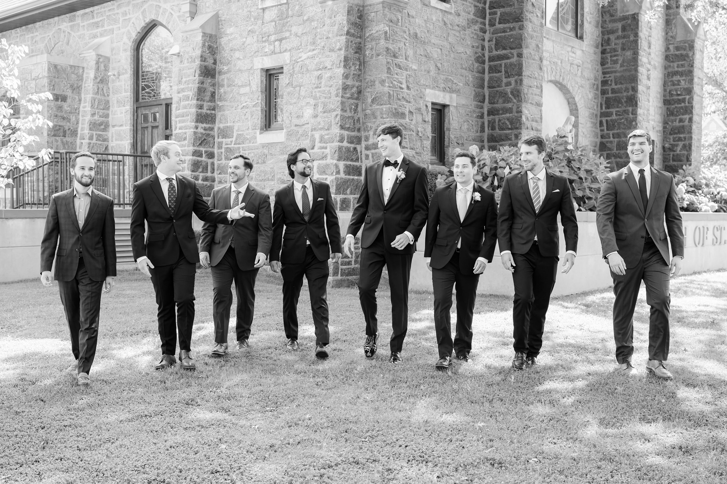 Groom walks confidently with his groomsmen outside a stone church, laughter and camaraderie capturing the joy and brotherhood of the wedding day.