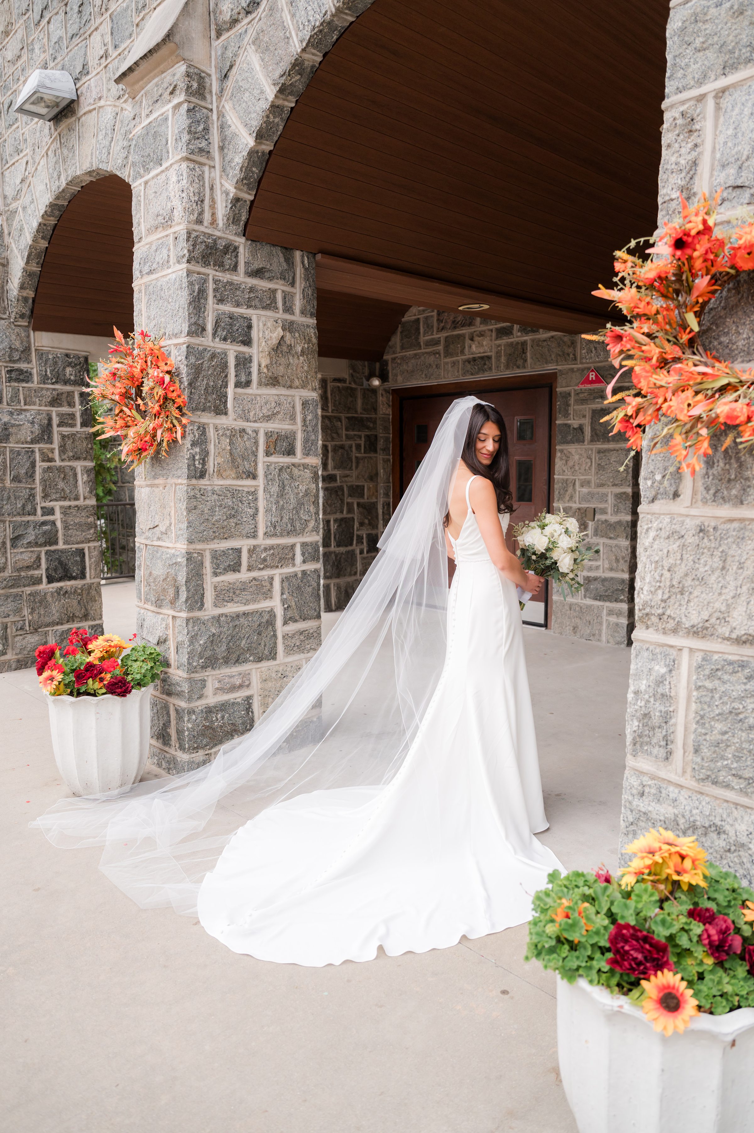 Bride pauses beneath stone arches, her veil flowing gently behind her as she turns with a quiet, joyful smile, holding her bouquet in a moment of calm before forever begins.