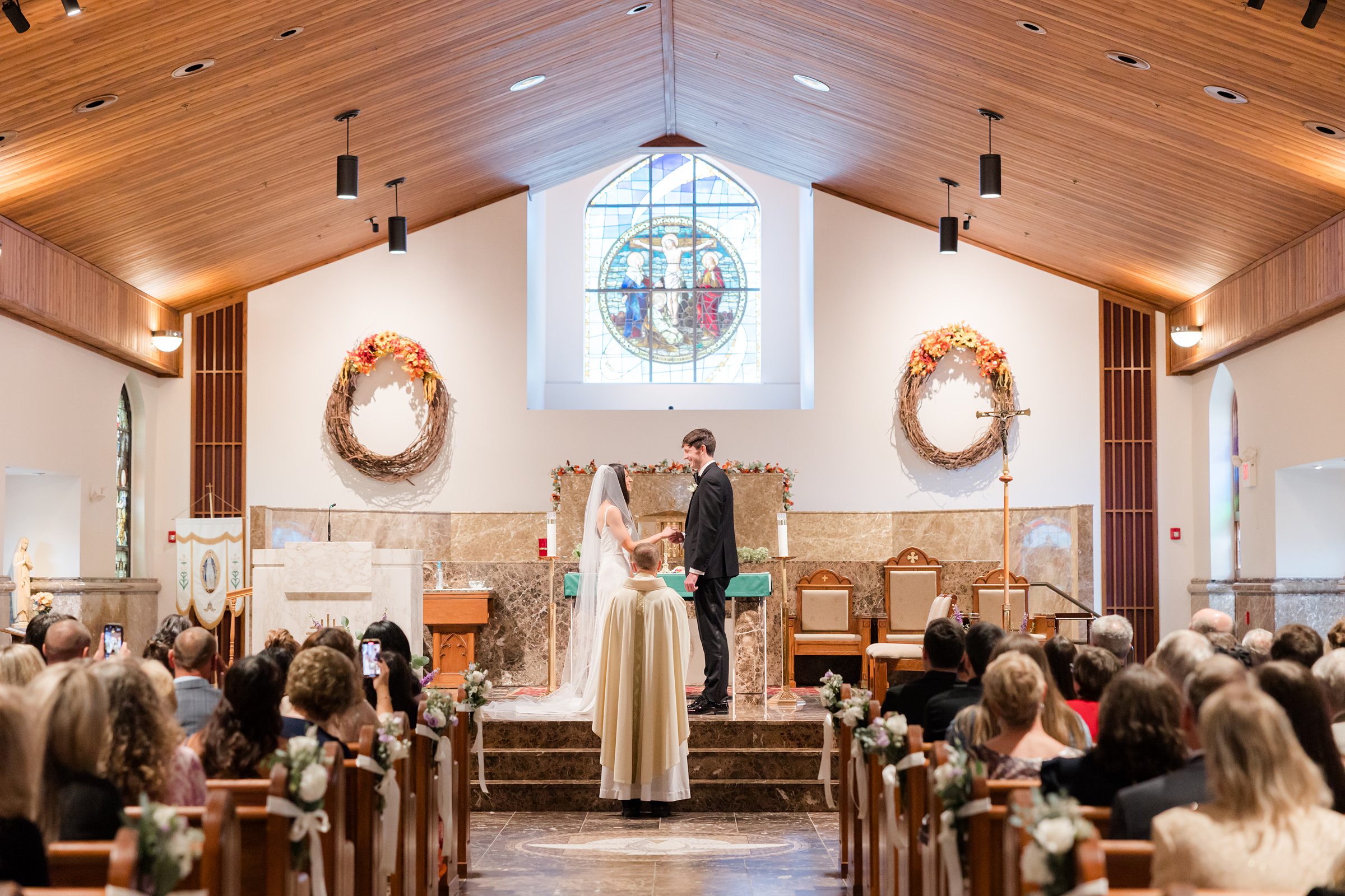 Bride and groom stand together at the altar, exchanging vows beneath soft light and stained glass, surrounded by loved ones witnessing the beginning of their forever.