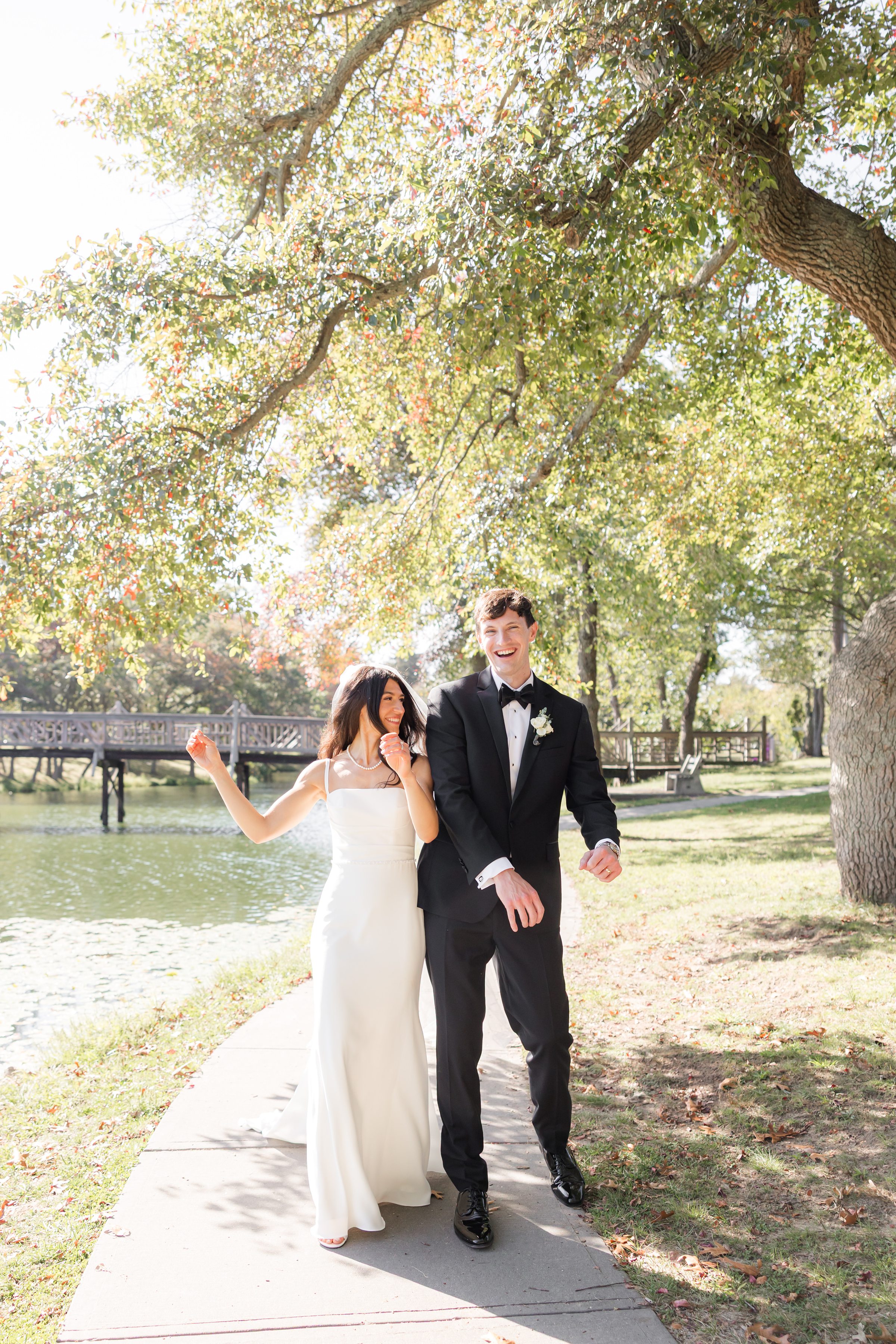 Bride and groom walk together beneath sunlit trees, laughing softly as they enjoy a playful, carefree moment.
