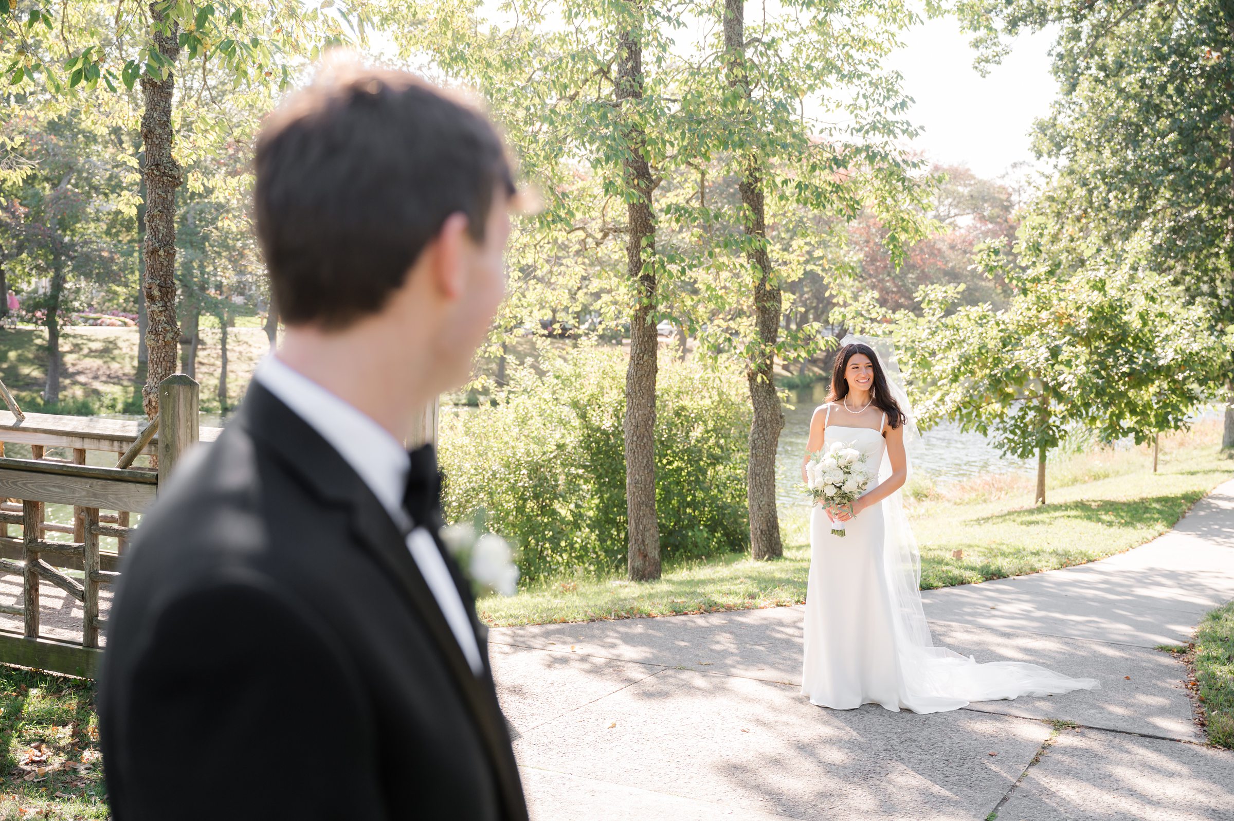 Groom turns to see his bride for the first time, her standing softly in the distance holding her bouquet, a moment filled with anticipation and love.