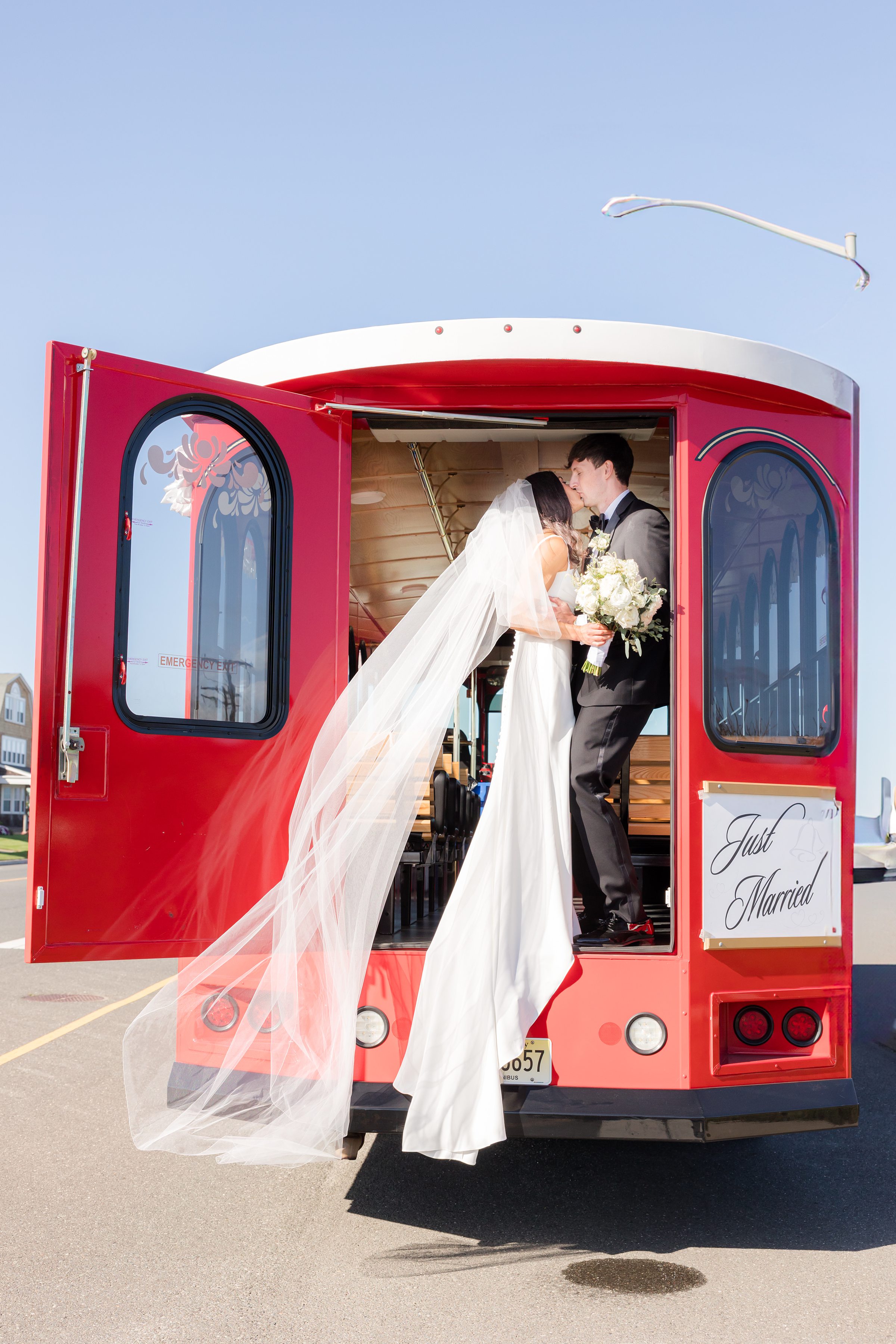 Bride and groom share a romantic kiss inside a bright red trolley, her flowing veil cascading behind her as they celebrate their just-married moment under a clear blue sky