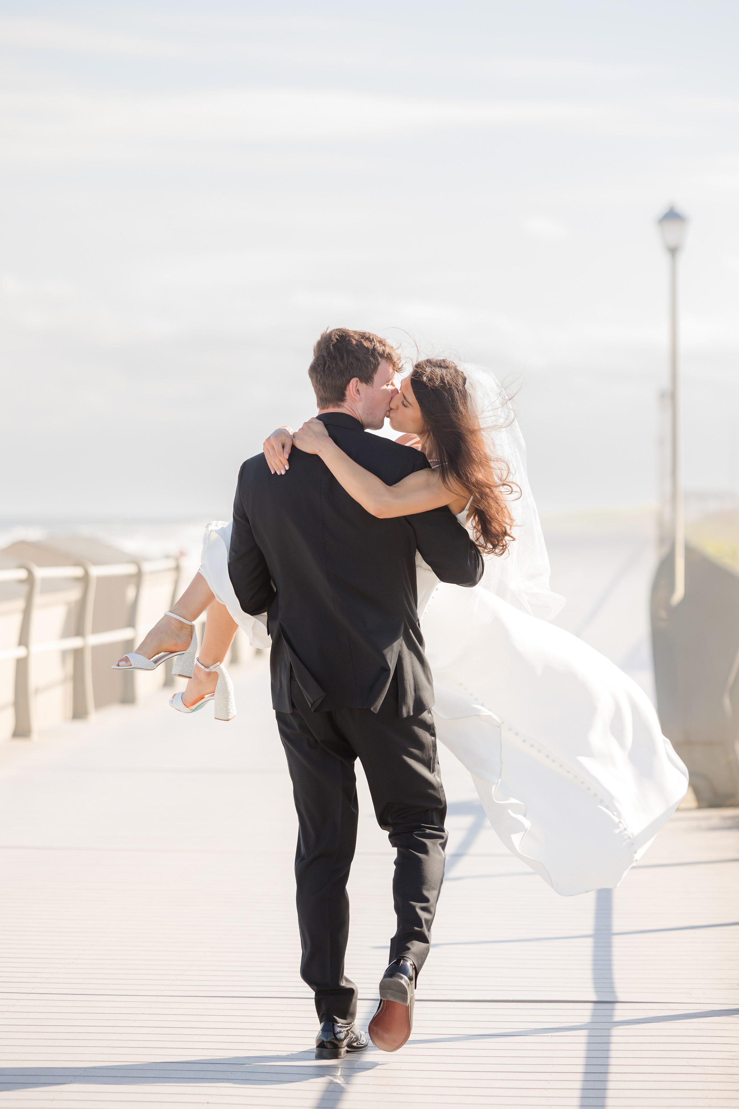 Groom lifts his bride into his arms for a kiss along the boardwalk, her dress flowing behind her in a cinematic, romantic moment