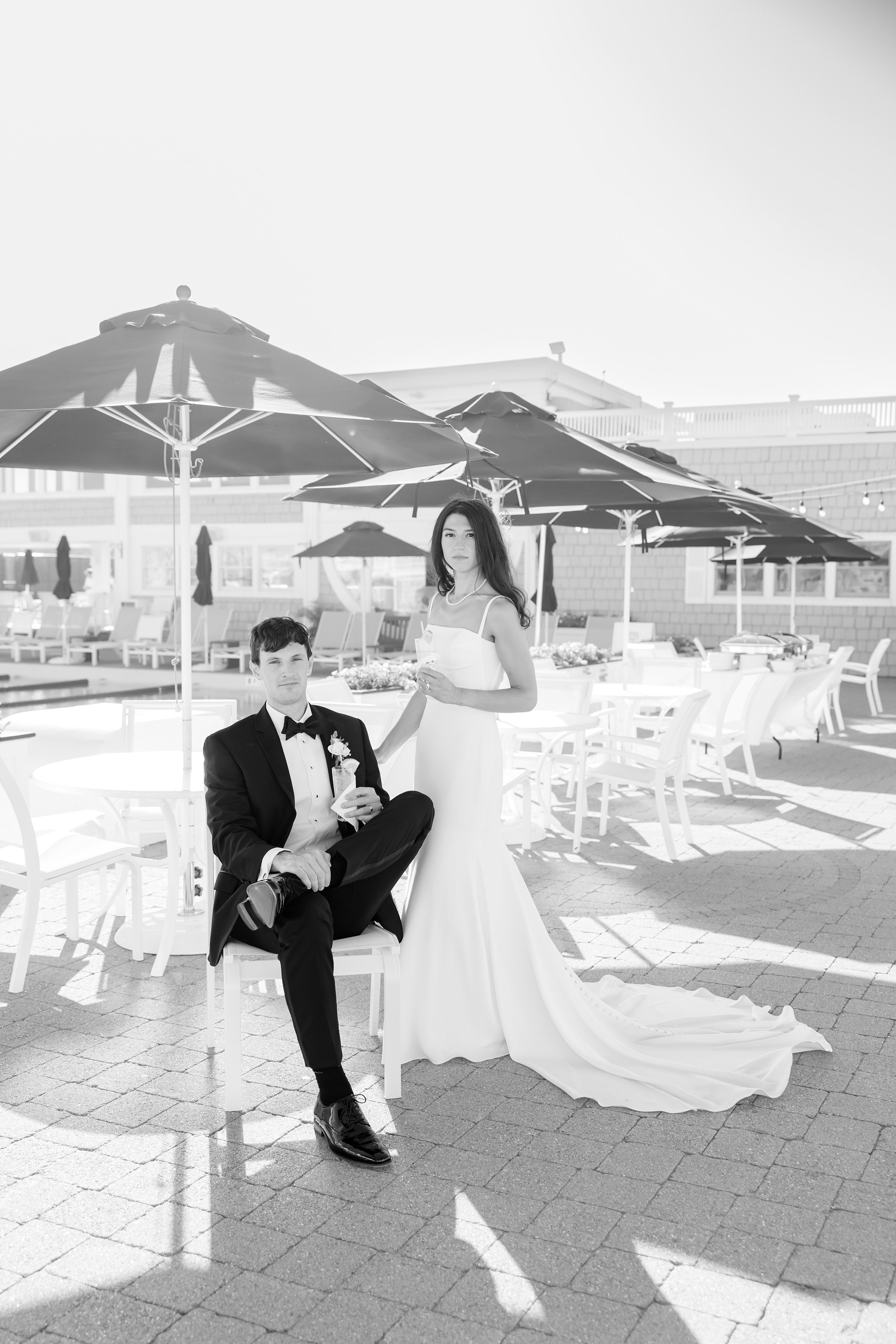 Bride stands gracefully beside her seated groom under sunlit umbrellas, both holding drinks and sharing a relaxed, elegant moment together by the pool on their wedding day