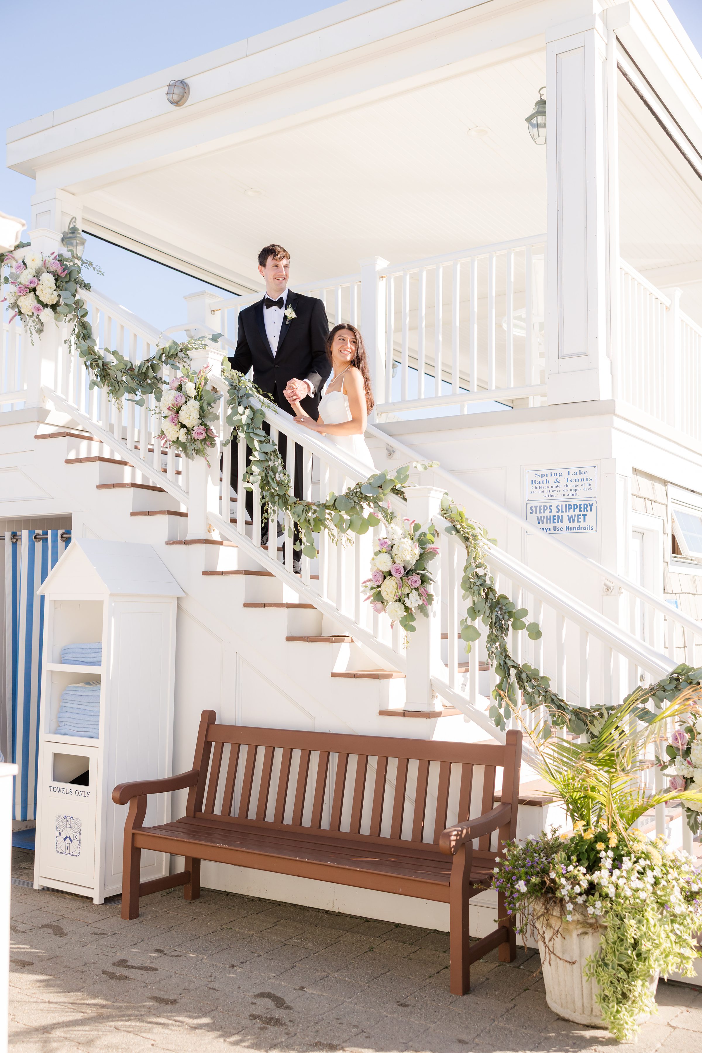Bride and groom pause on a sunlit white staircase adorned with soft florals and greenery, smiling together as they take in the joy of their wedding day.