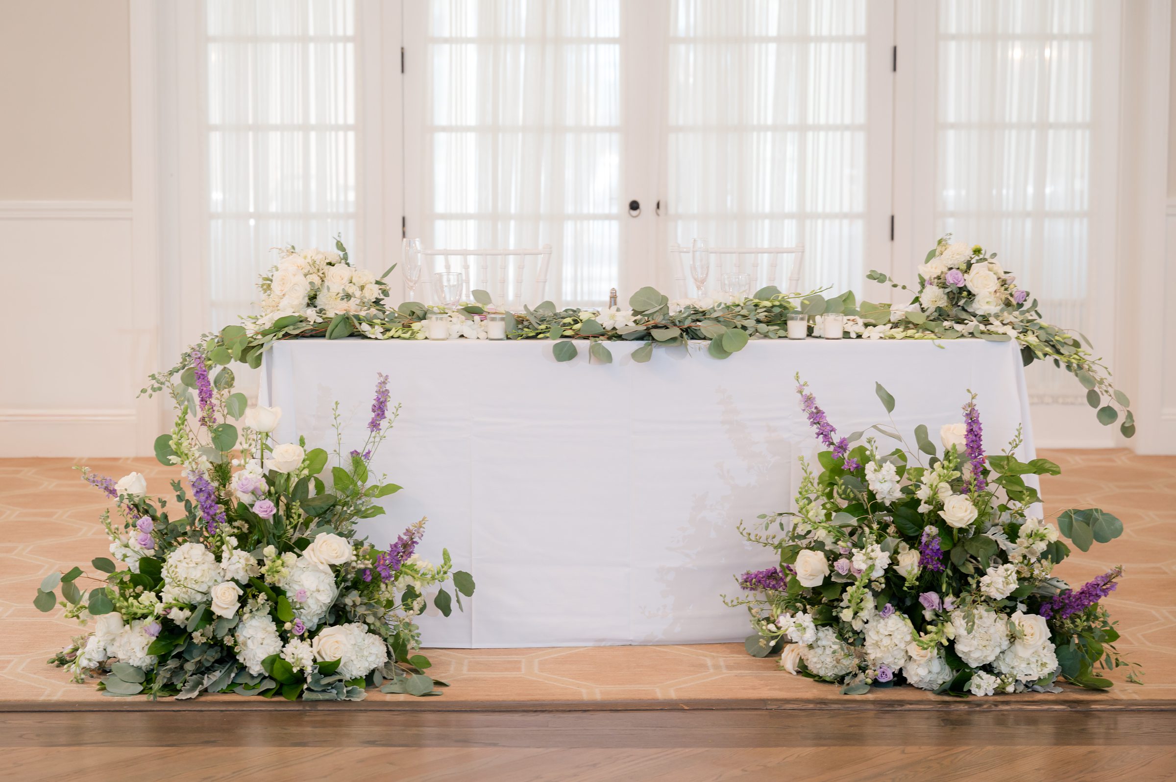 Beautifully styled sweetheart table adorned with lush white and lavender floral arrangements and cascading greenery, set against soft, light-filled doors for an elegant and romantic reception backdrop.