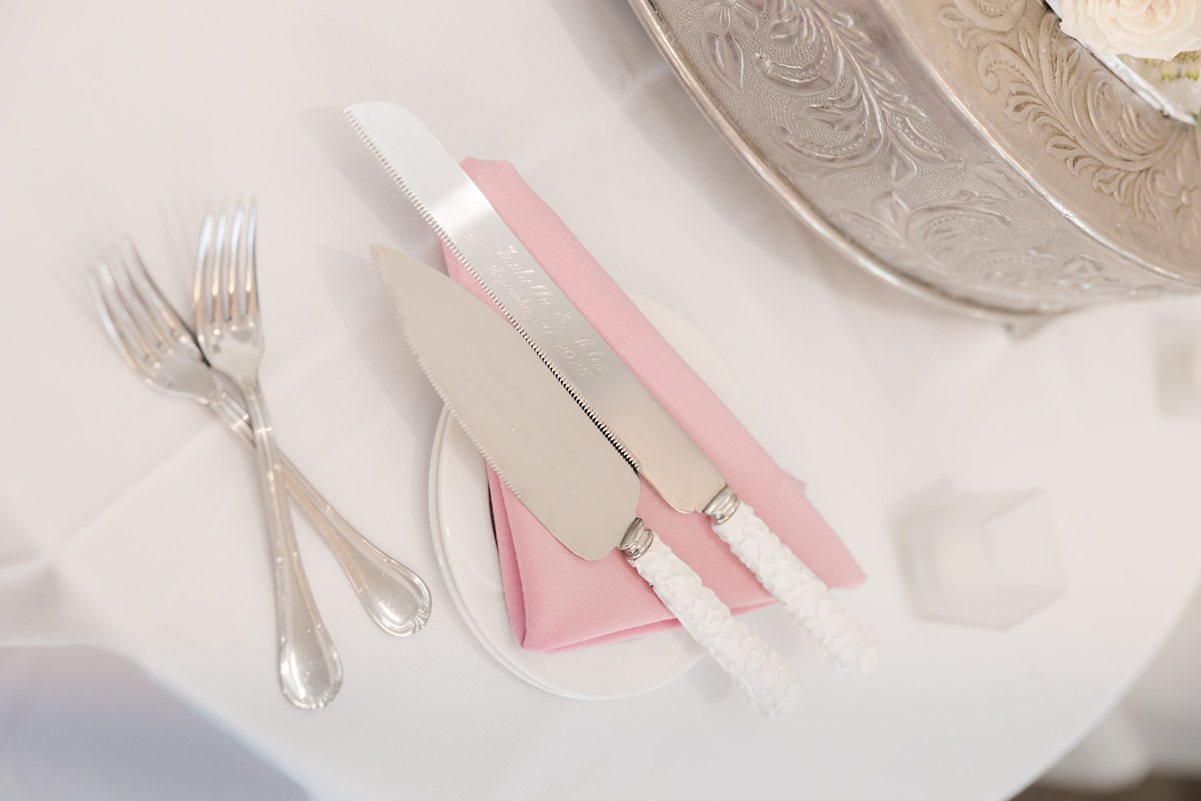 Elegant wedding cake cutting set rests atop a soft pink napkin beside polished silverware, capturing a delicate and refined detail of the reception décor.