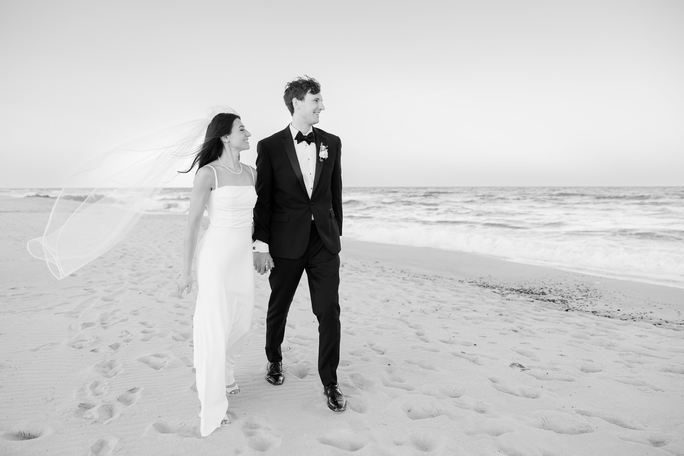 Bride and groom walk together along the shoreline, her veil flowing in the ocean breeze as they share a quiet, romantic moment by the sea.