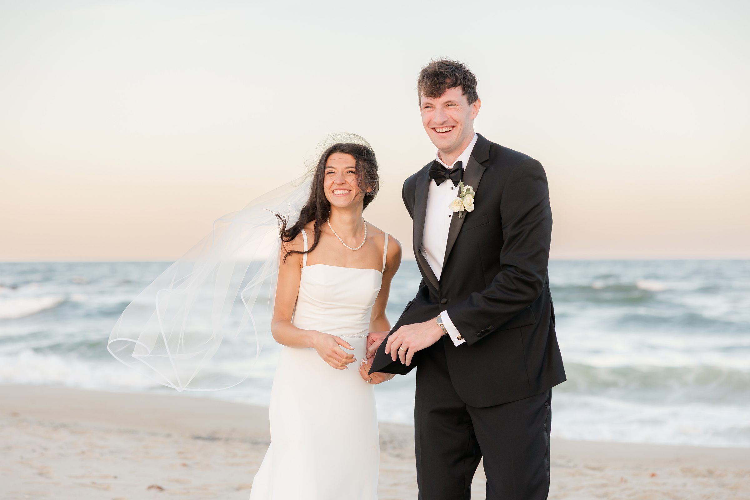 Bride and groom stand together on the beach, smiling brightly as the ocean stretches behind them under soft evening light.