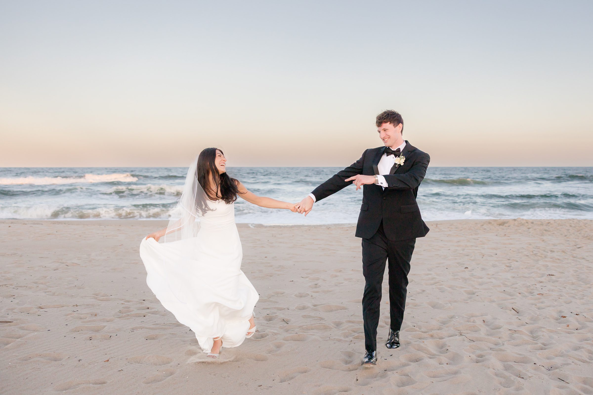 Bride and groom hold hands and laugh while walking along the beach at sunset, waves rolling gently behind them.