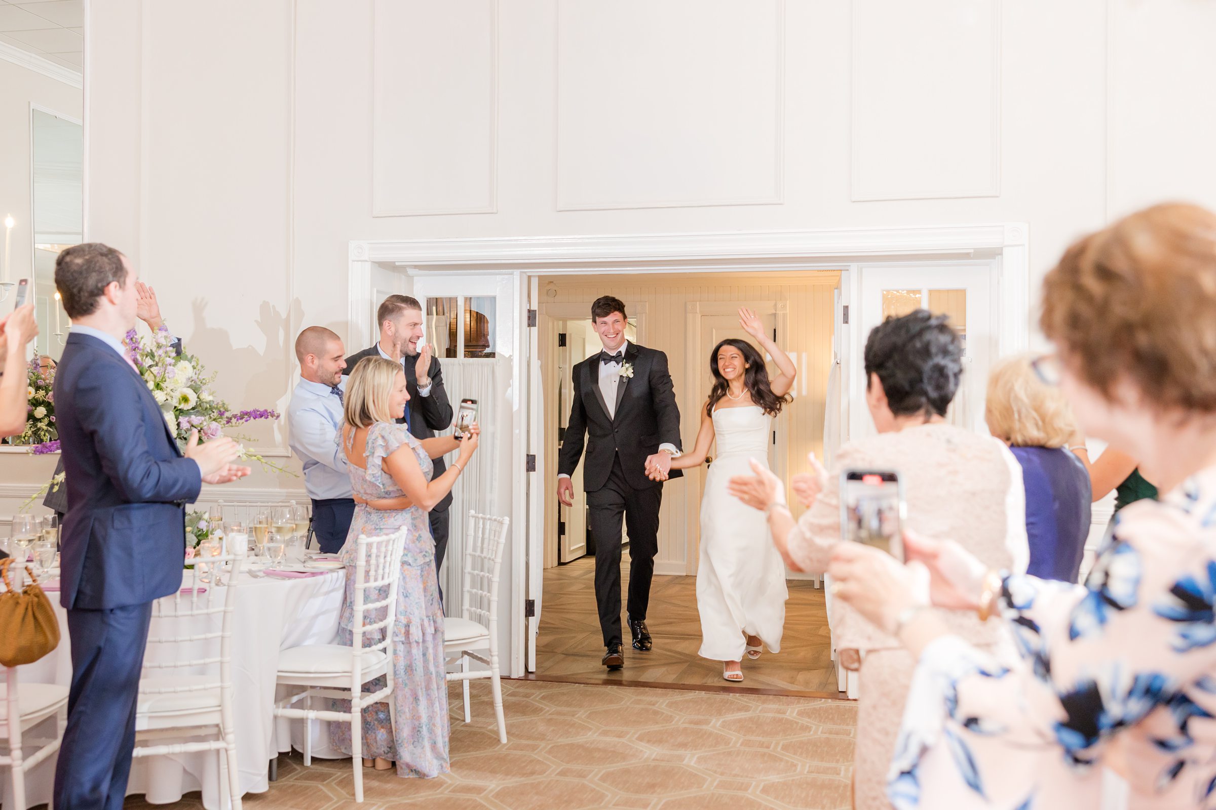 Newlyweds enter their reception together, smiling and waving as guests stand and applaud their arrival.