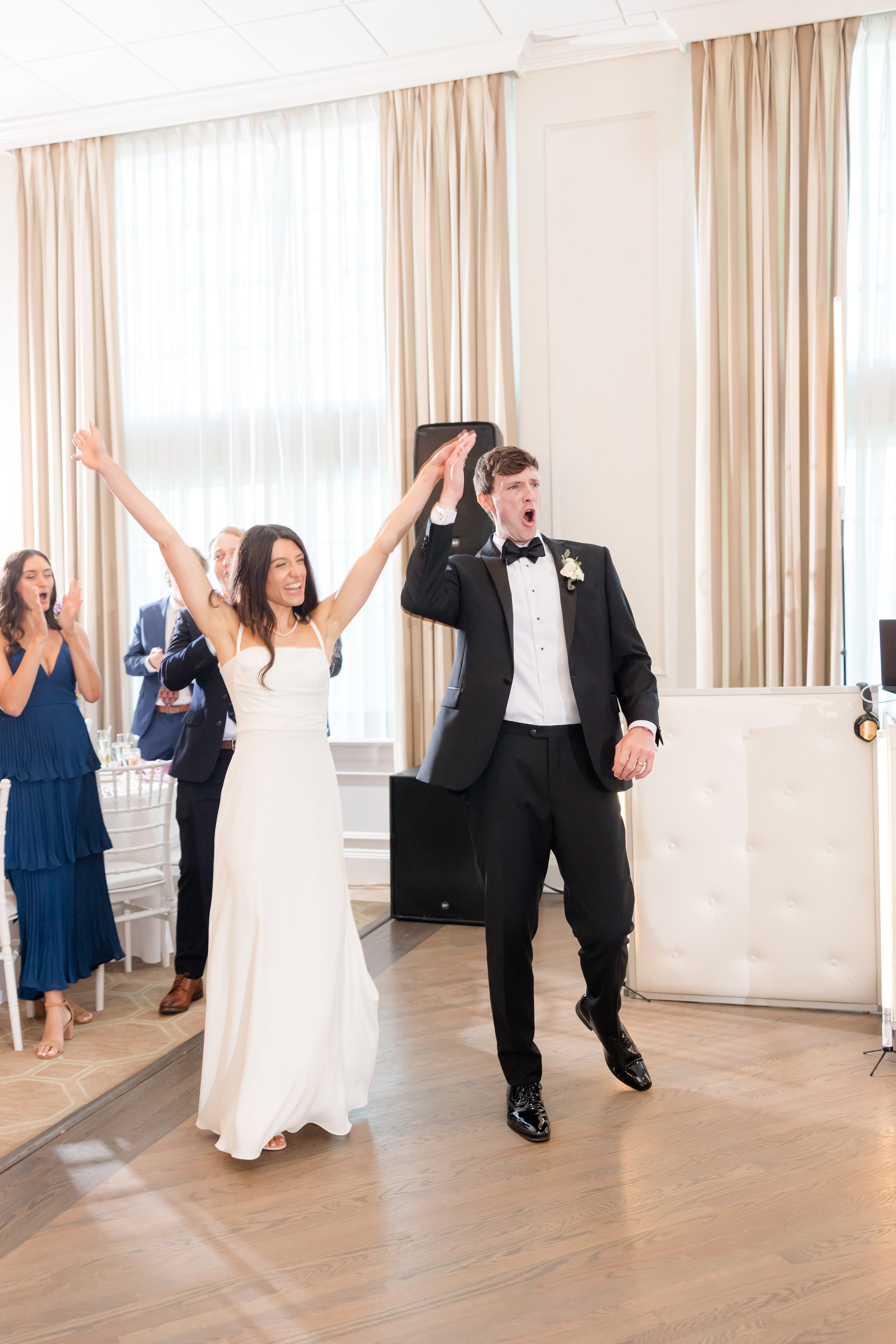 Bride and groom raise their hands in celebration as they enter the reception, smiling and full of excitement while guests applaud behind them.