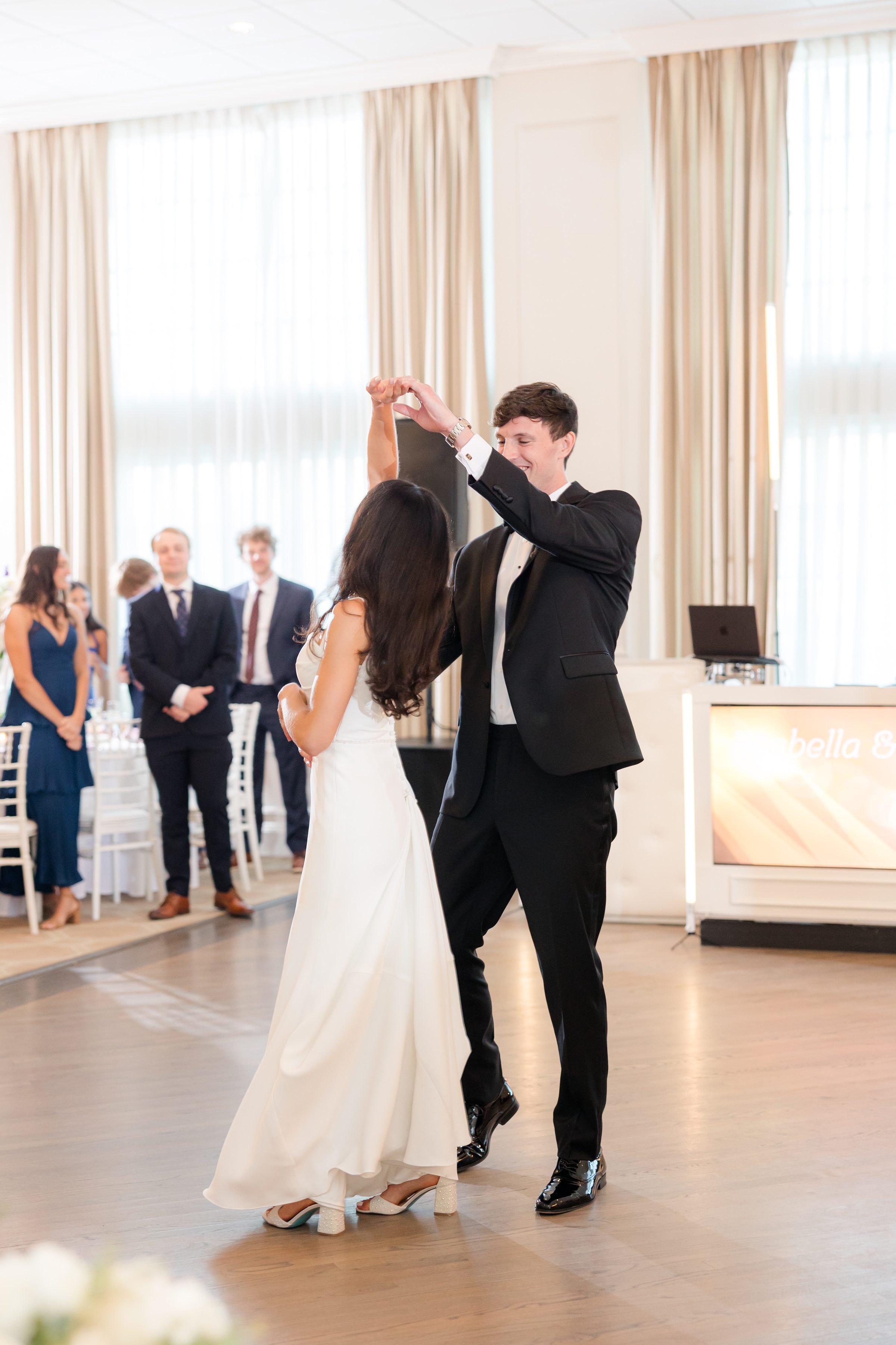 Groom twirls his bride across the dance floor as they share a joyful first dance, surrounded by softly lit reception decor and watching guests.