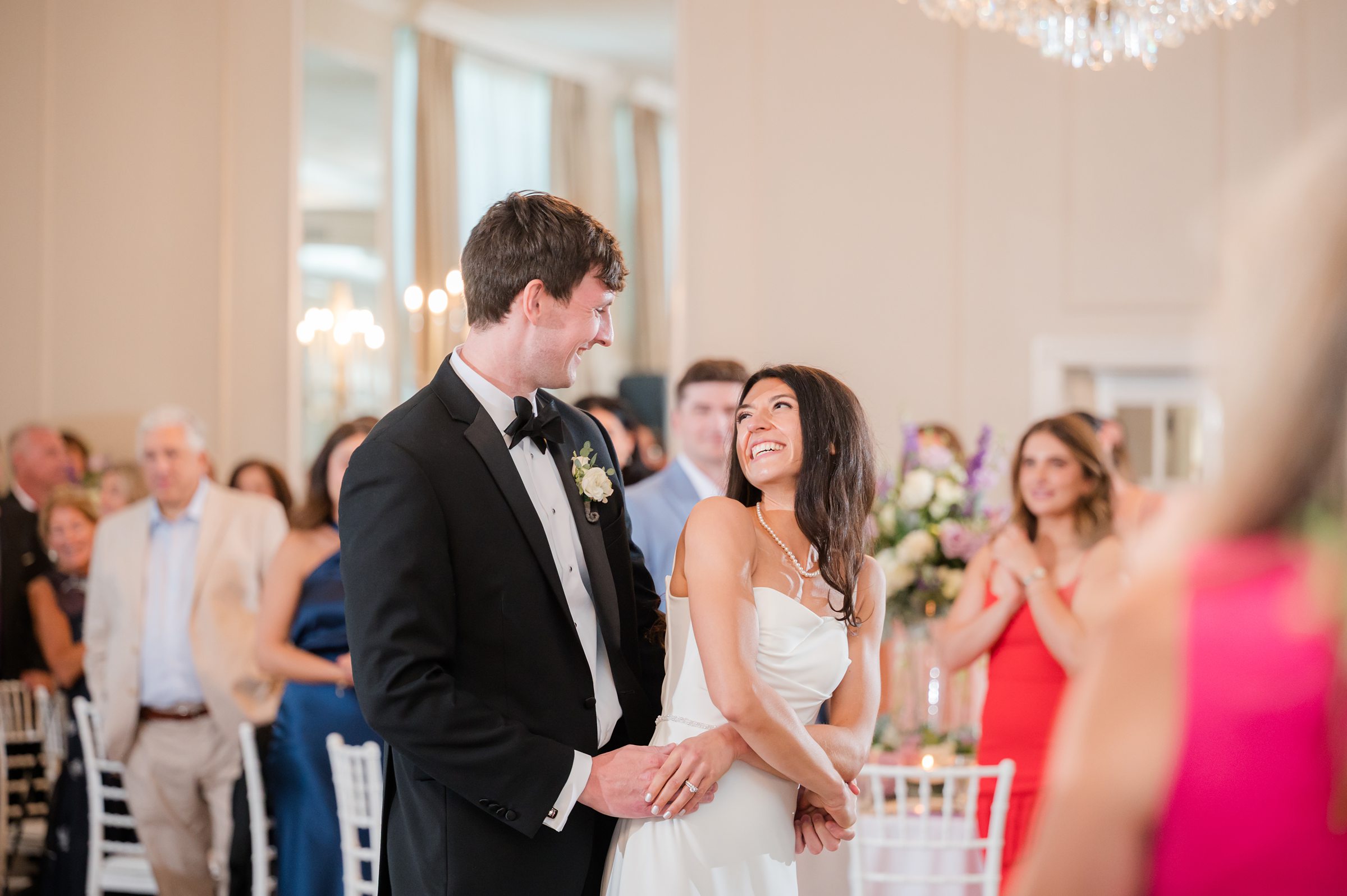 Bride and groom smile at each other during their first dance, surrounded by guests watching and celebrating in a softly lit ballroom