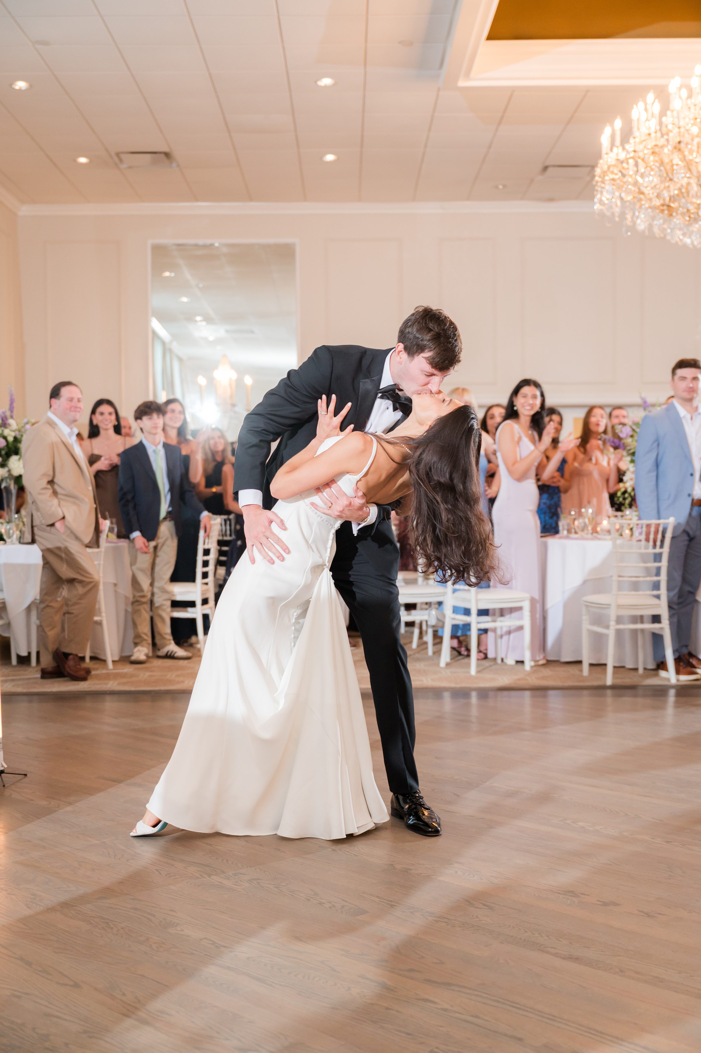 Groom dips his bride into a kiss on the dance floor as guests watch and cheer in an elegant ballroom.