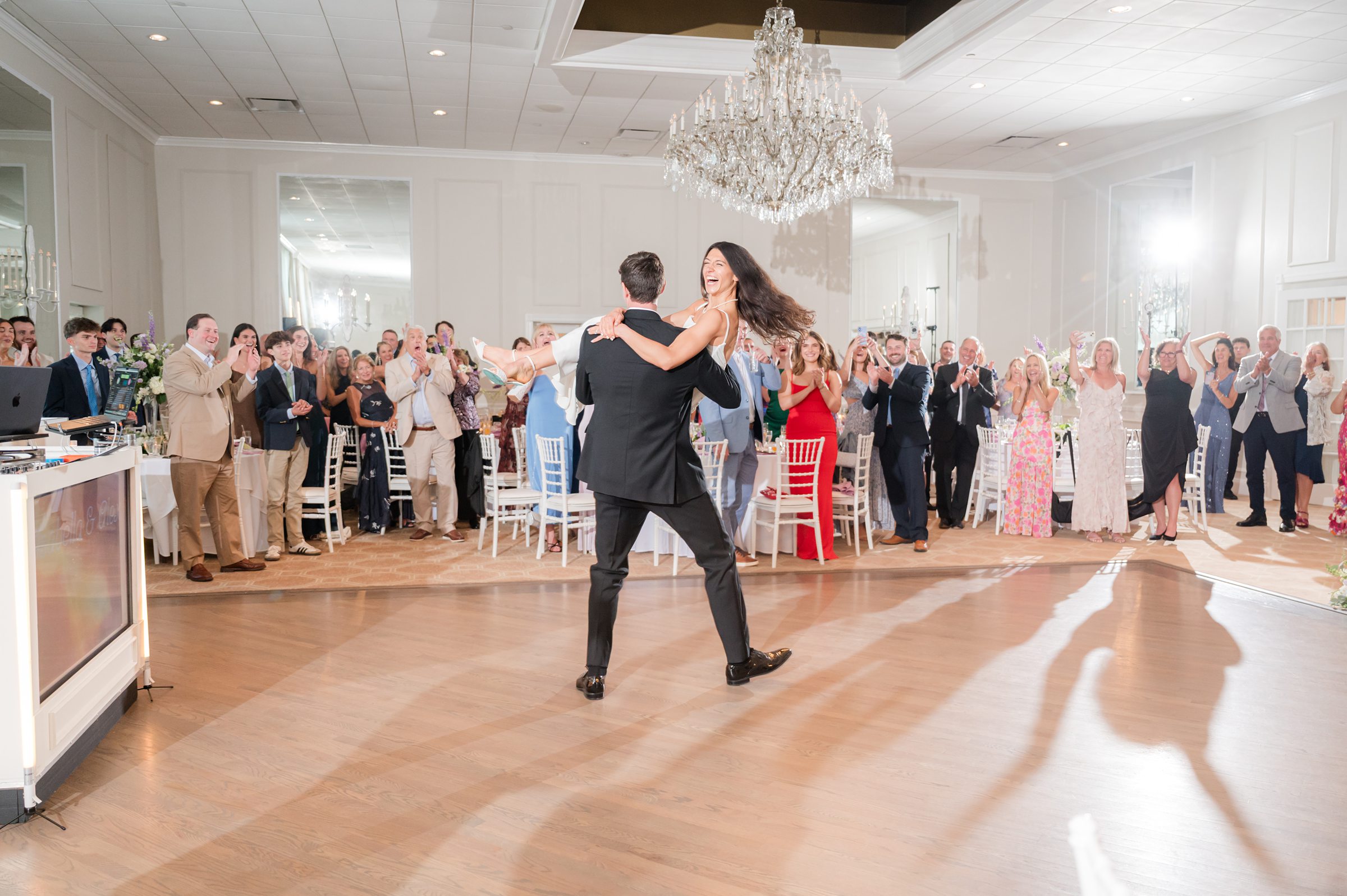 Groom lifts his bride during their first dance as guests surround them, clapping and cheering in a bright, elegant ballroom