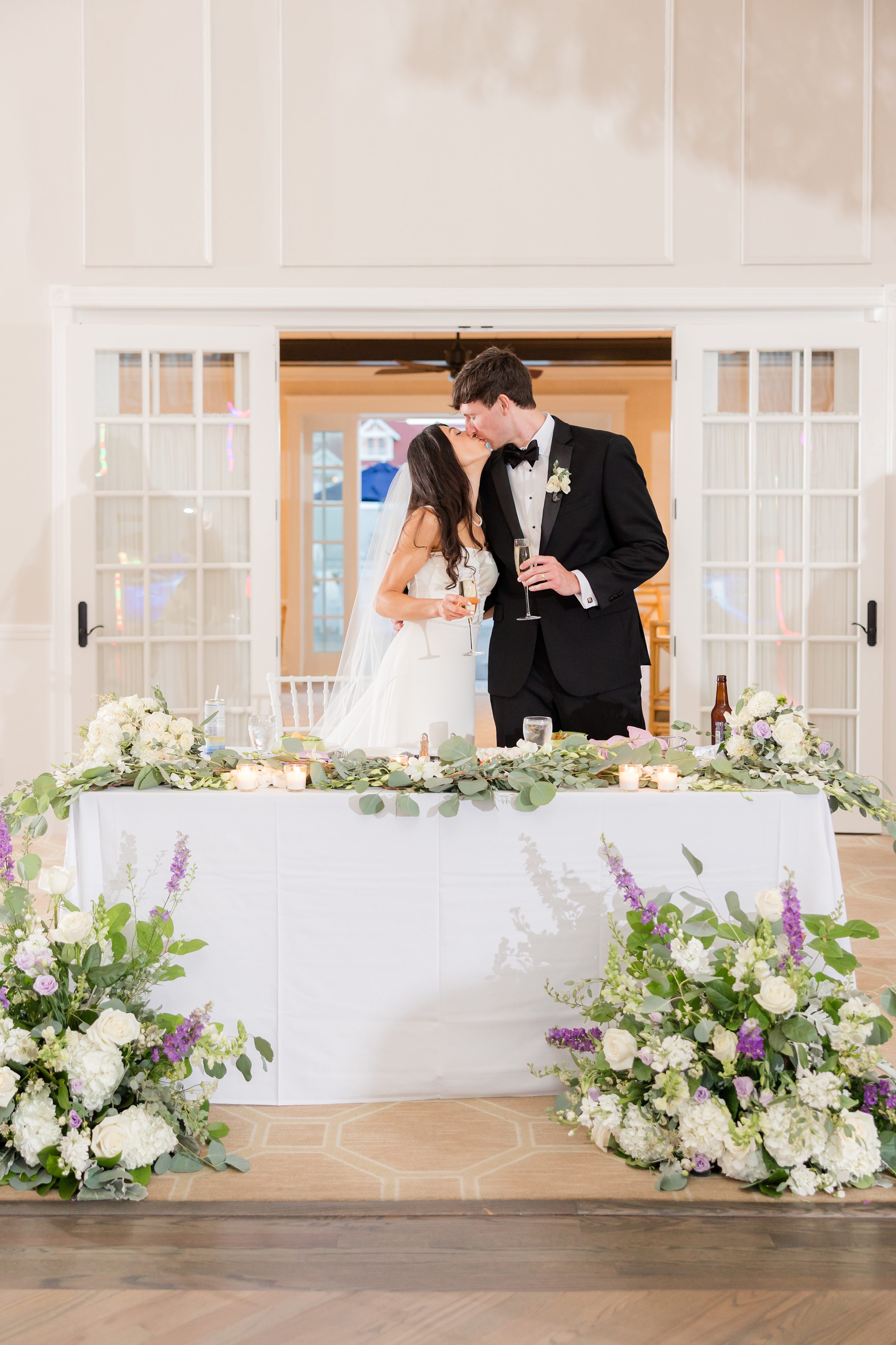 Bride and groom share a kiss at their sweetheart table, holding champagne glasses and surrounded by candles and soft floral arrangements