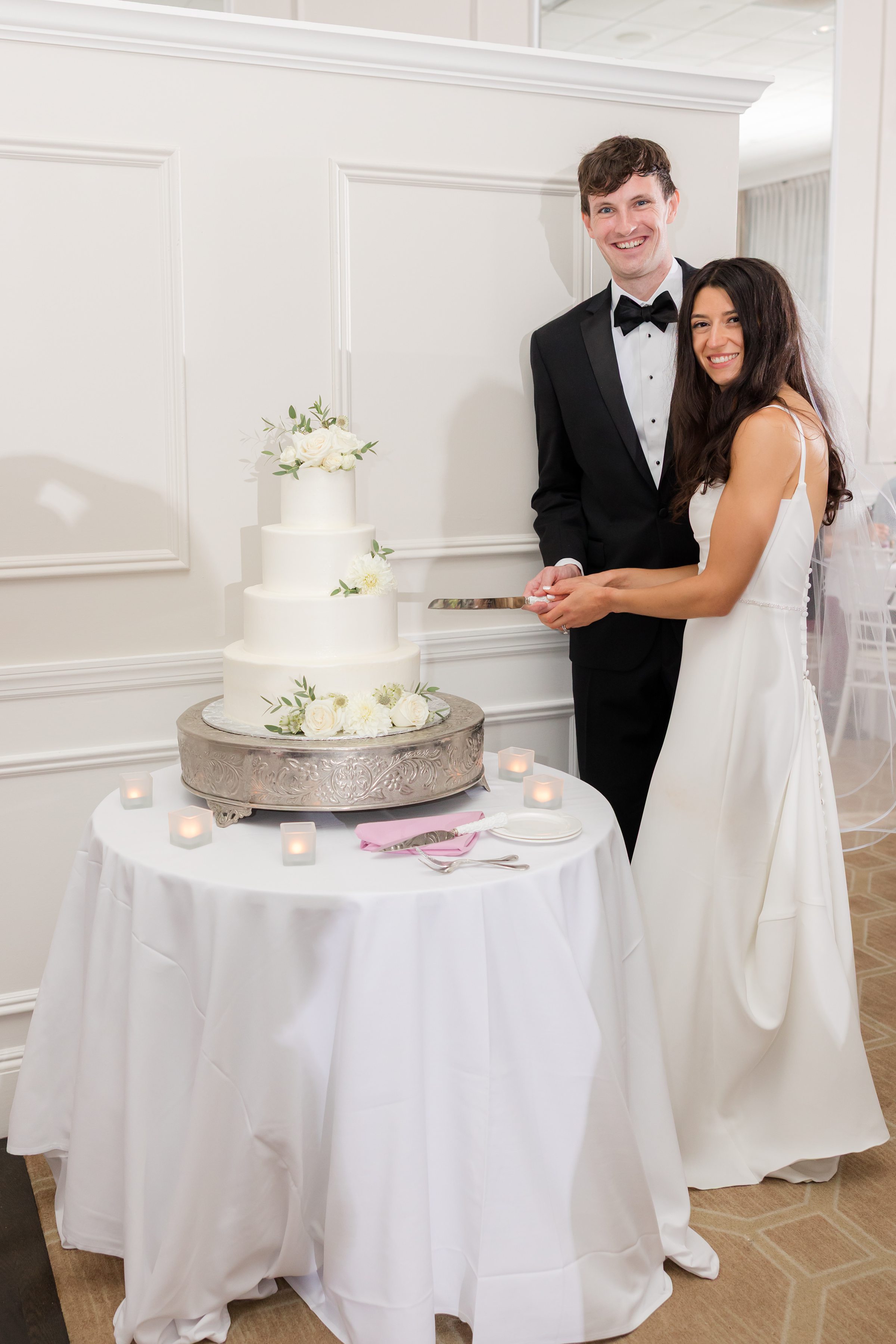 Newlyweds stand close together, smiling warmly as they cut their floral wedding cake in a softly lit reception space.