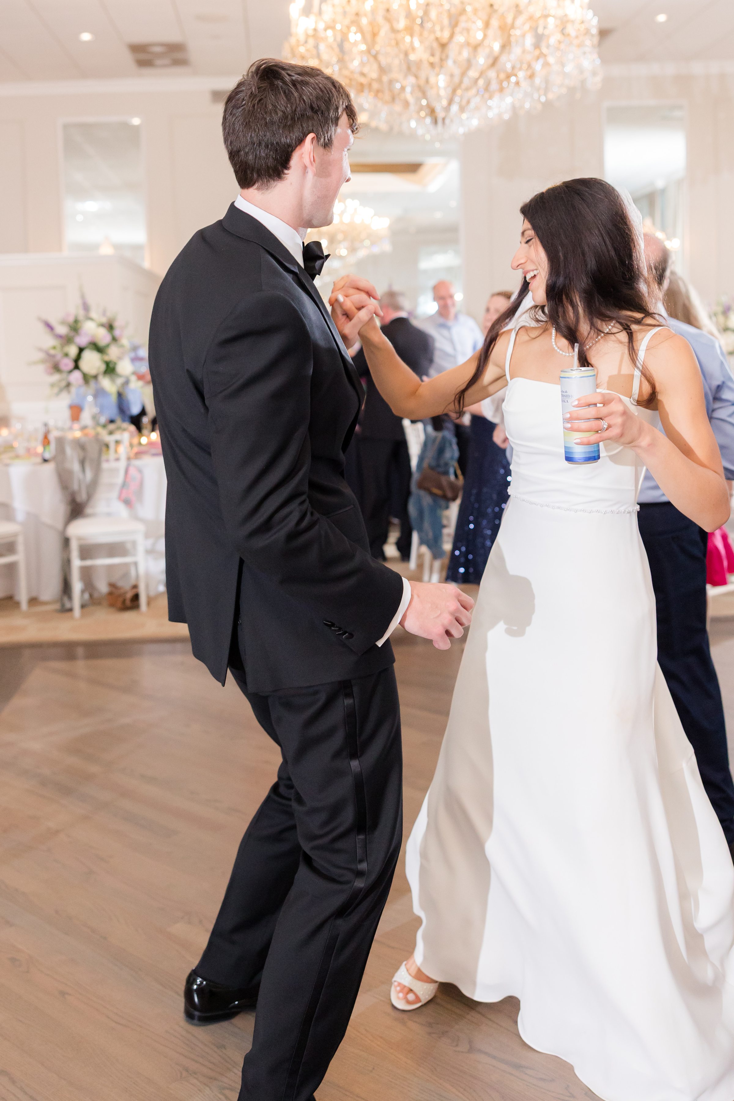 Bride and groom laugh and dance together on the reception floor, sharing a joyful moment beneath a sparkling chandelier.