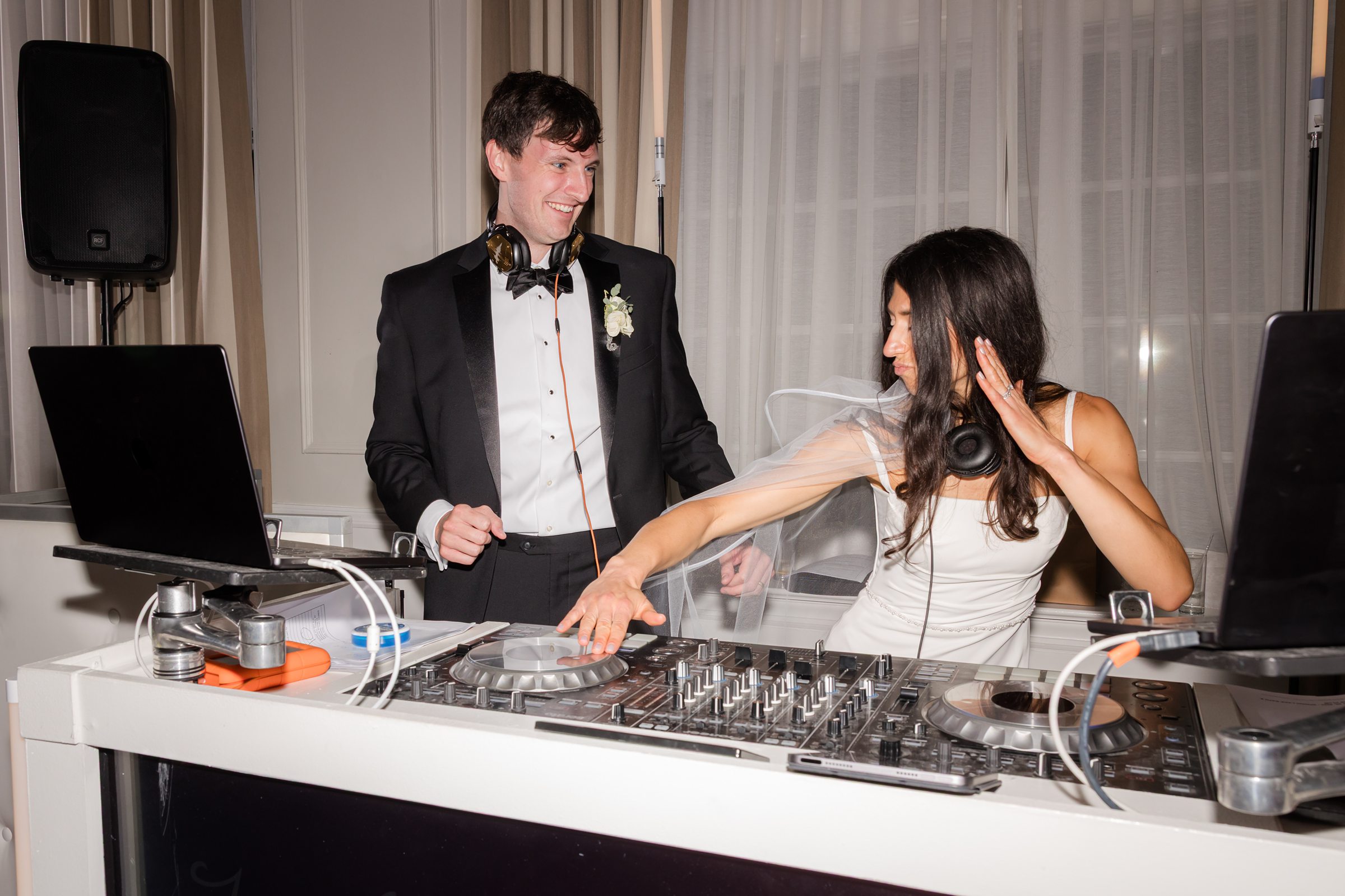 Groom smiles warmly as he watches his bride DJ and dance, sharing a playful and unforgettable reception moment.