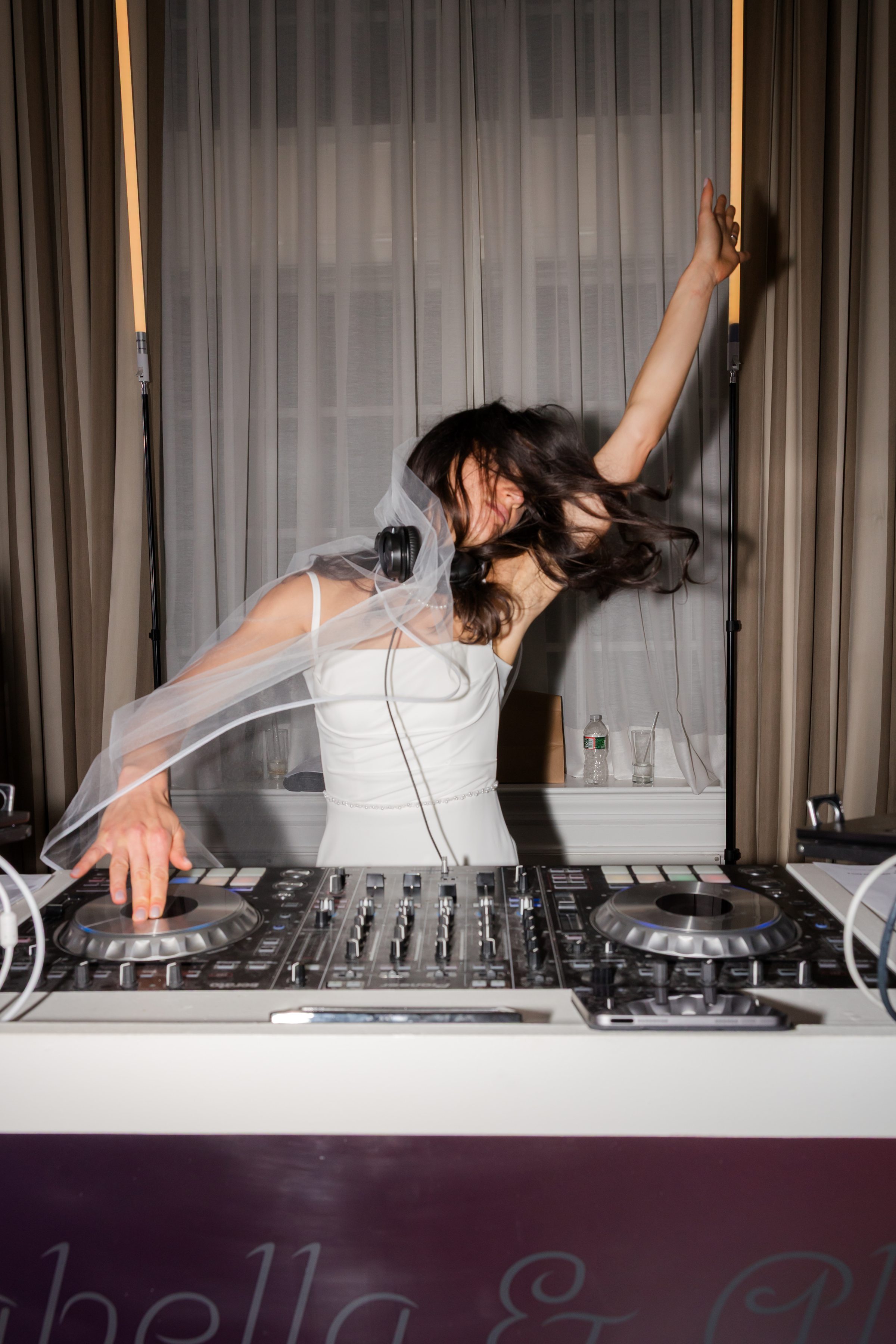 Bride joyfully dances at the DJ booth, veil flowing and hair in motion, fully immersed in the celebration