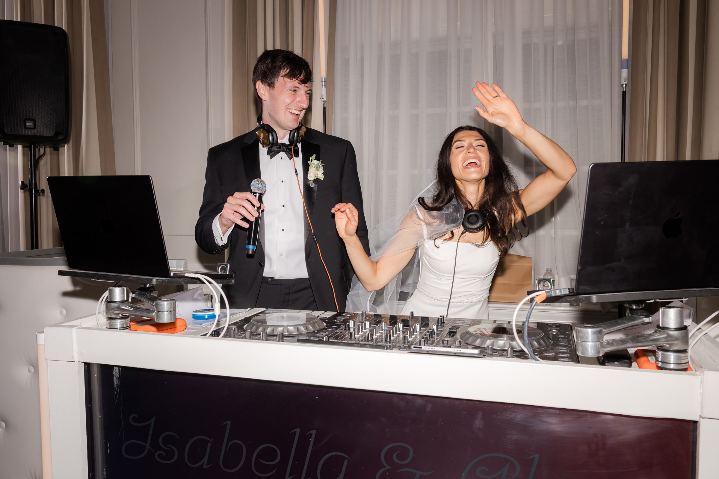Bride and groom celebrate joyfully behind the DJ booth, laughing and dancing together as they embrace the excitement of their wedding night