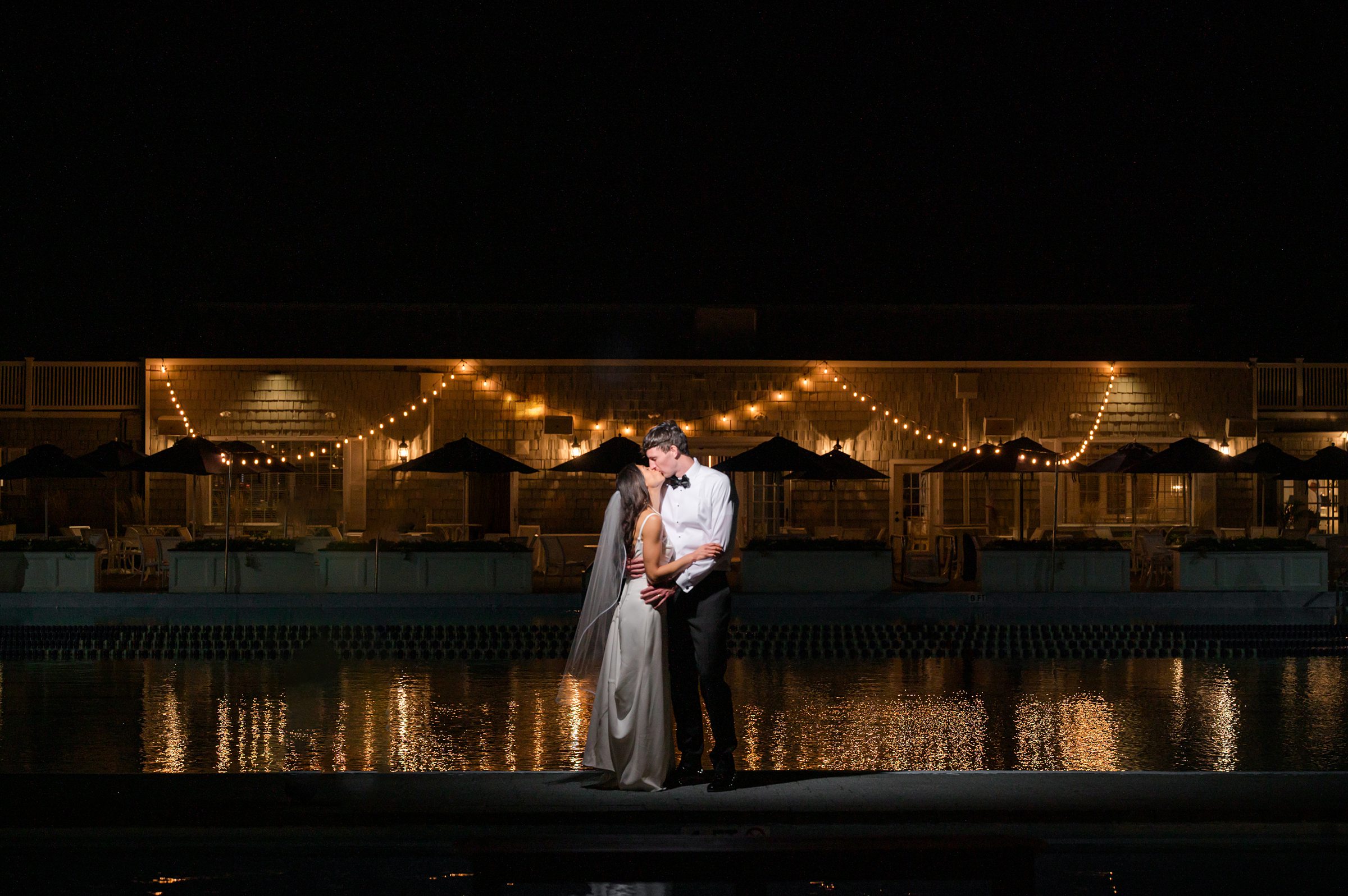 Bride and groom share a quiet embrace and kiss by the water at night, surrounded by warm string lights reflecting across the pool