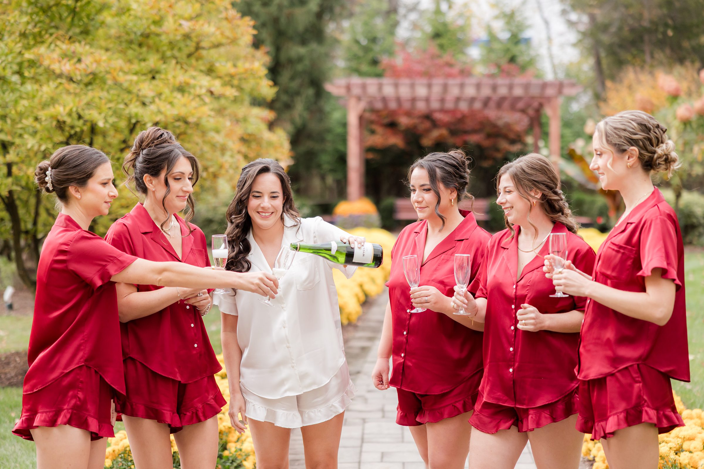Bride and bridesmaids in matching red outfits toasting with champagne outdoors.