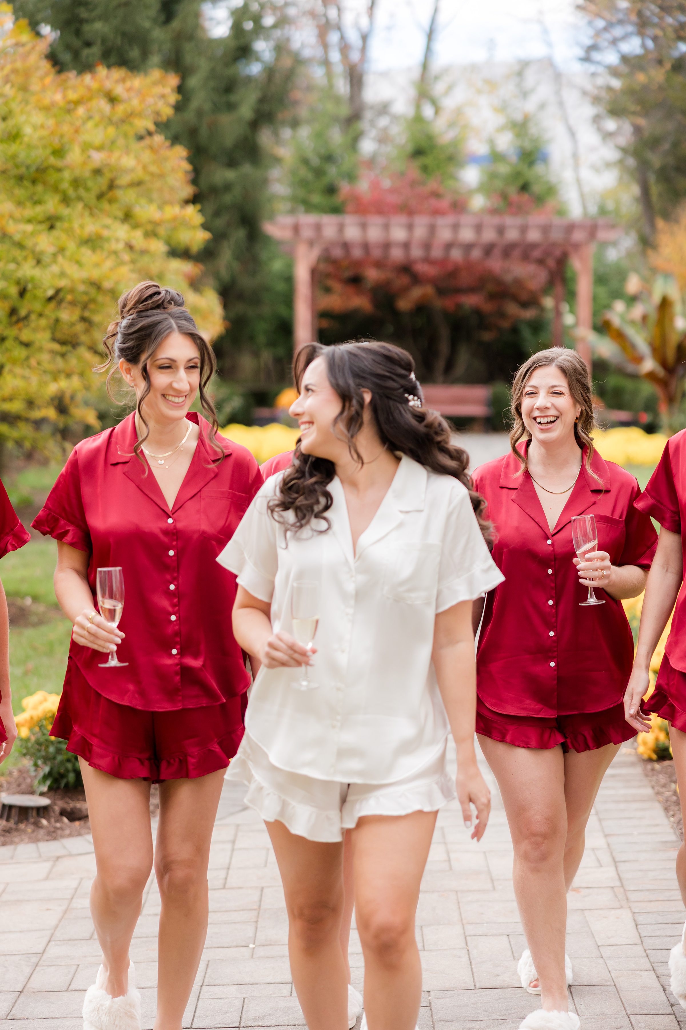 Bride walking with bridesmaids in red outfits, smiling and holding champagne glasses in a garden.
