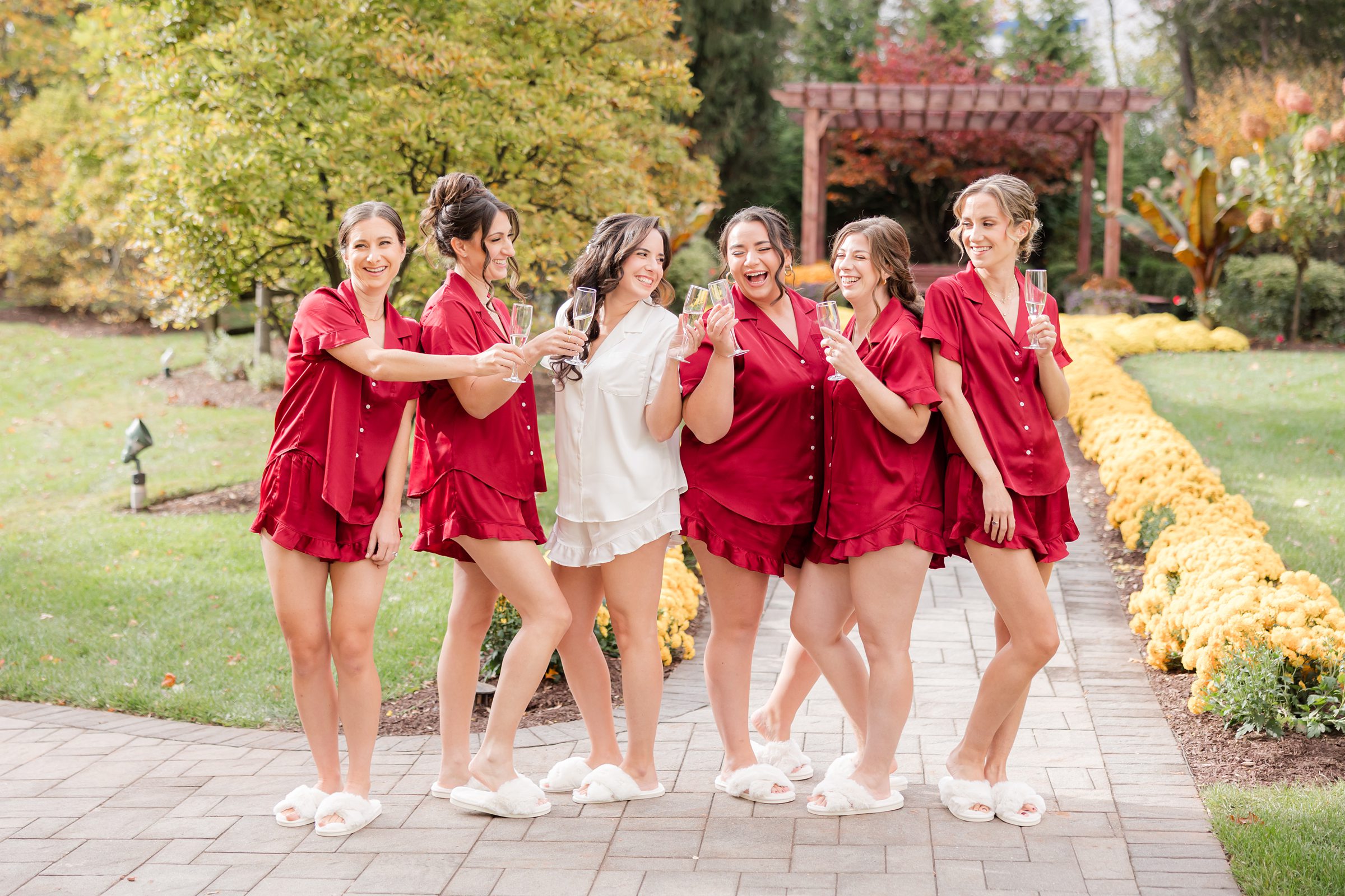 Bride and bridesmaids in red pajamas laughing and posing with champagne in a garden.