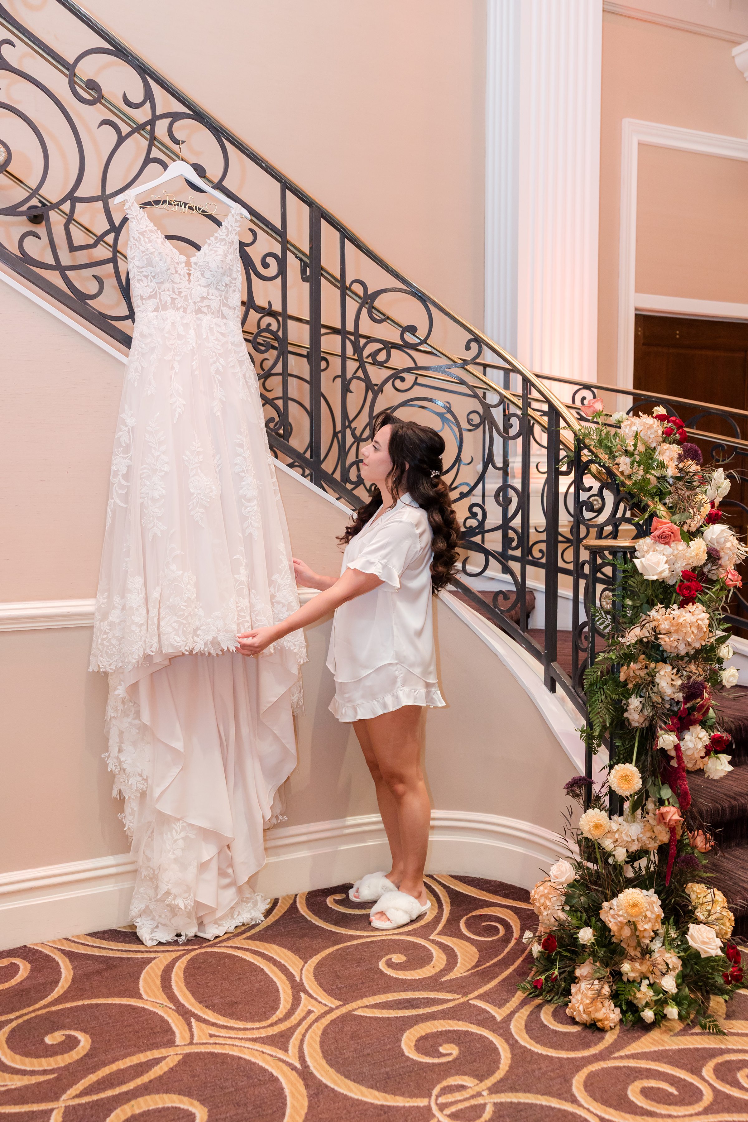 Bride admiring her wedding dress hanging on an elegant staircase with floral decor.