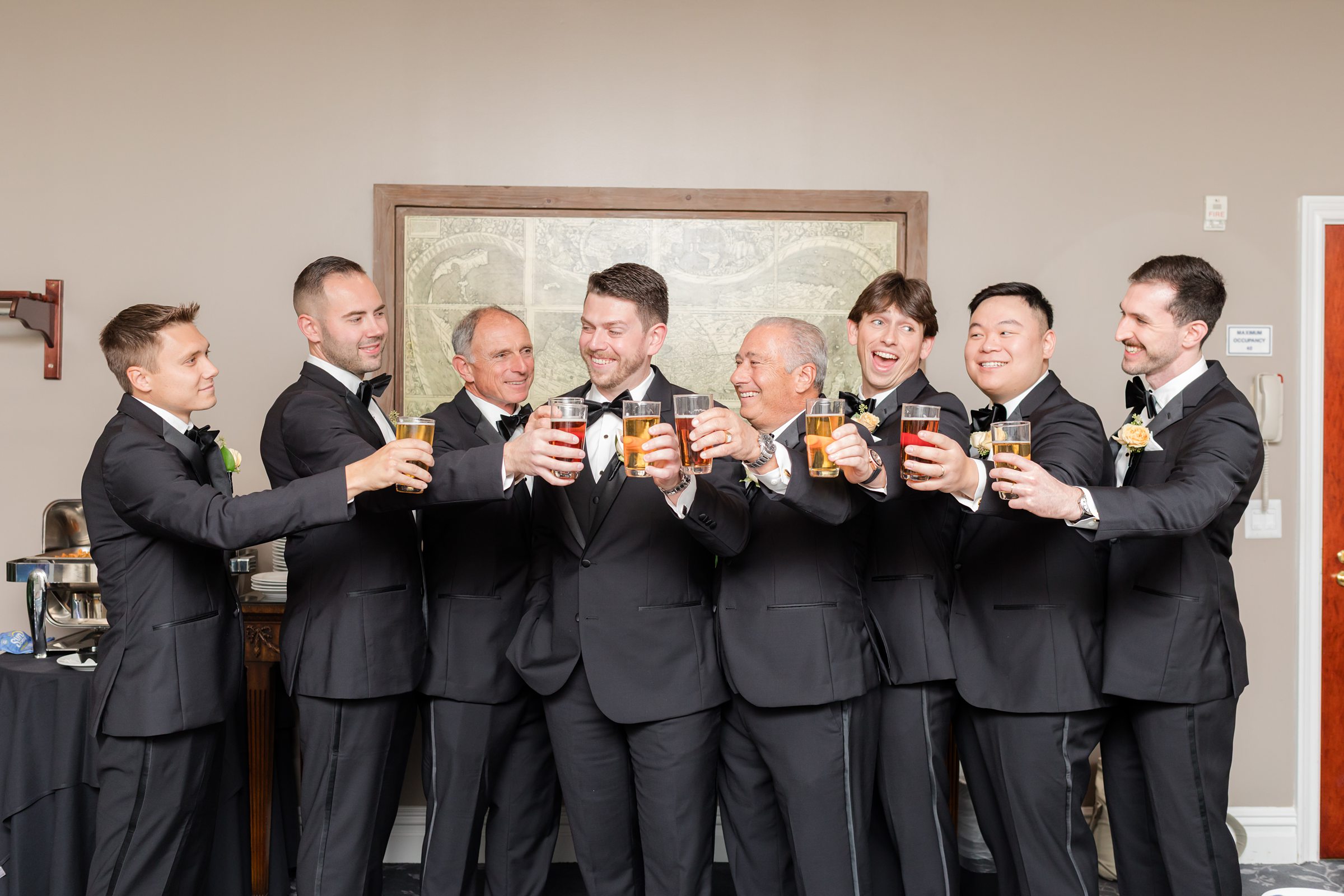 Groom and groomsmen in black tuxedos raising a toast and smiling together before the wedding.