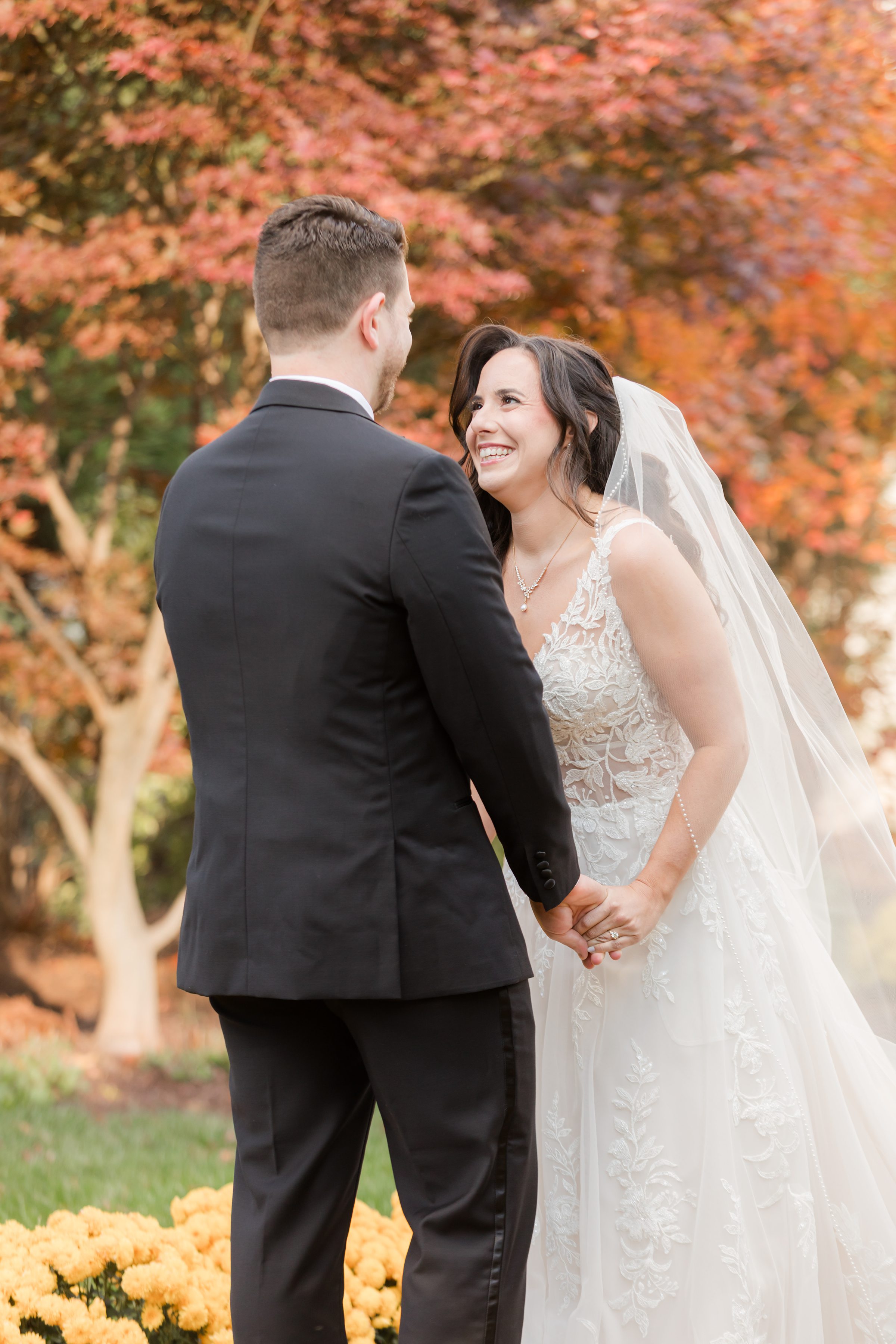 Bride and groom holding hands and smiling at each other during their first look in an autumn garden.
