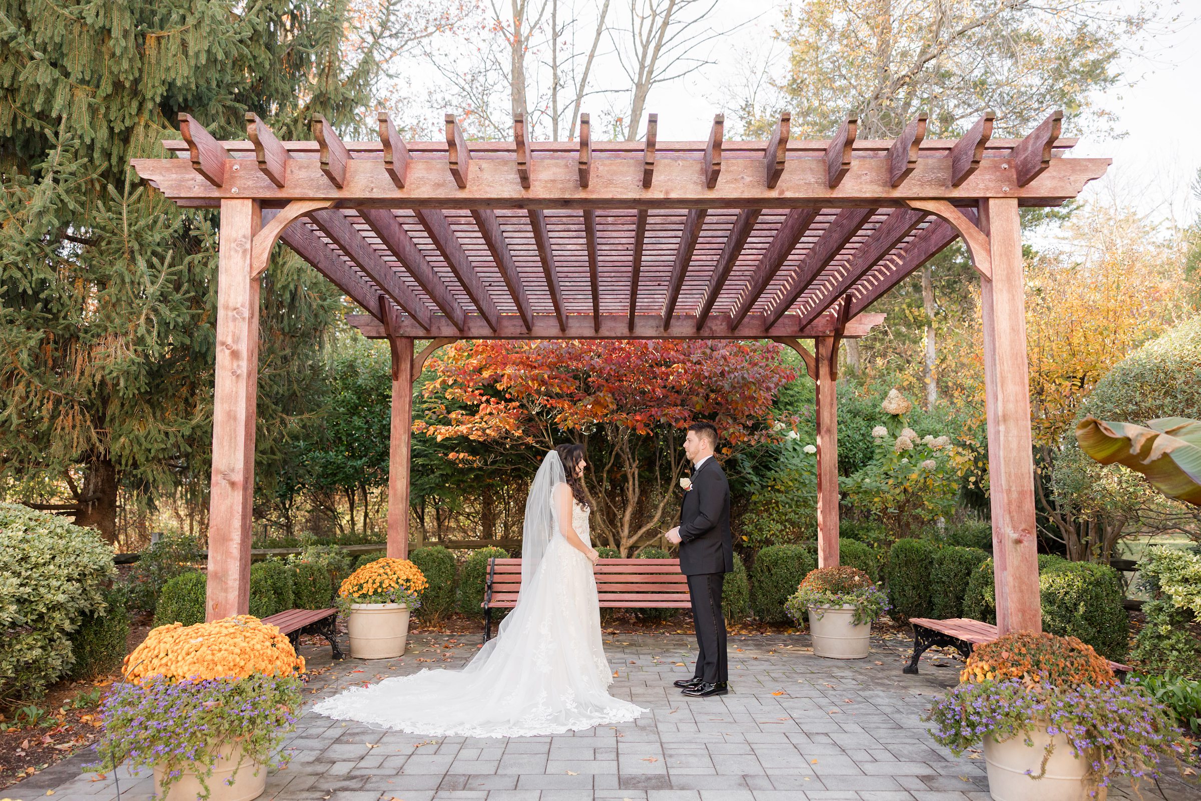 Beneath a rustic wooden pergola, the bride and groom stand facing each other in a quiet, intimate moment, surrounded by soft autumn colors as they soak in the beginning of their forever together.