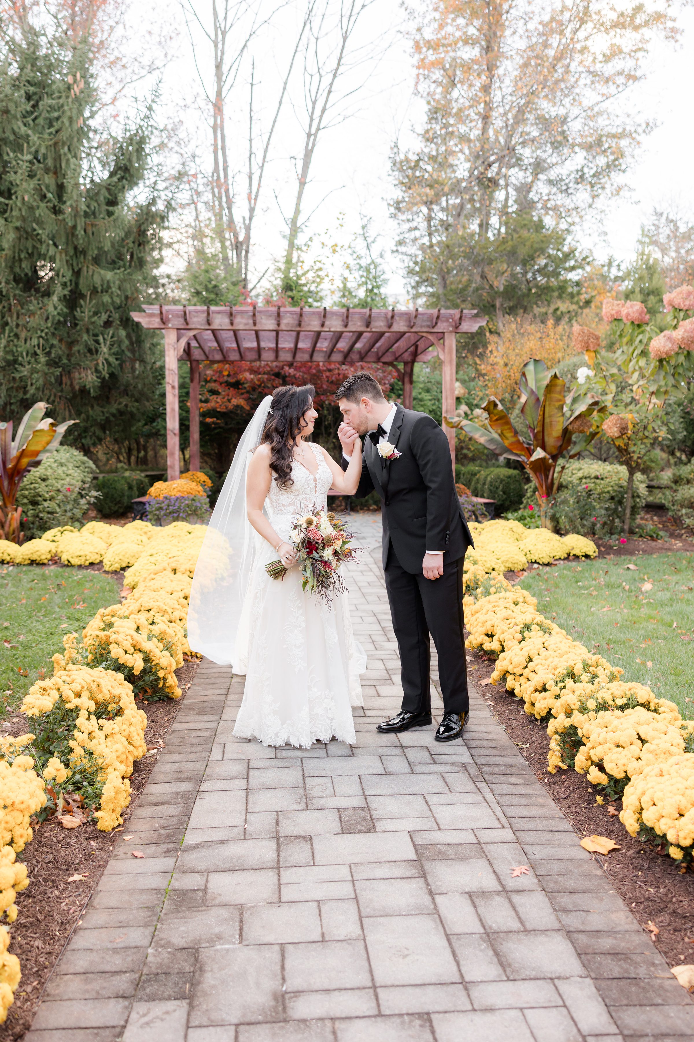 Groom lifting the bride’s hand for a kiss beneath a wooden pergola surrounded by lush greenery and fall flowers.