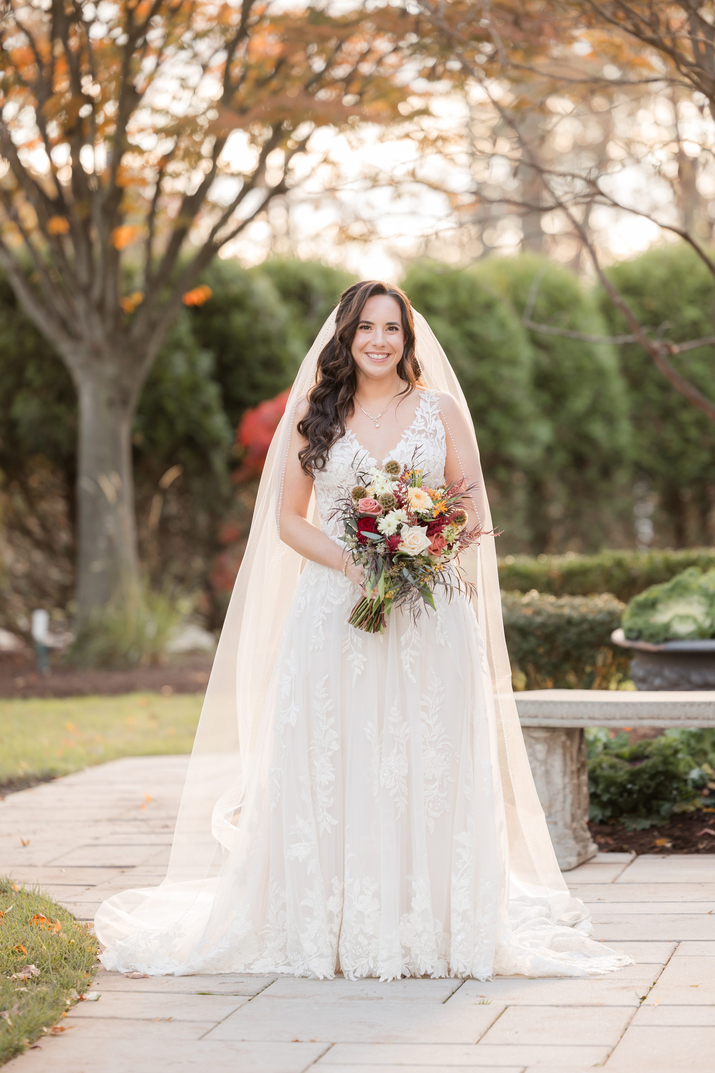 Bride standing alone holding her bouquet, smiling warmly in a garden of soft greenery and fall tones.