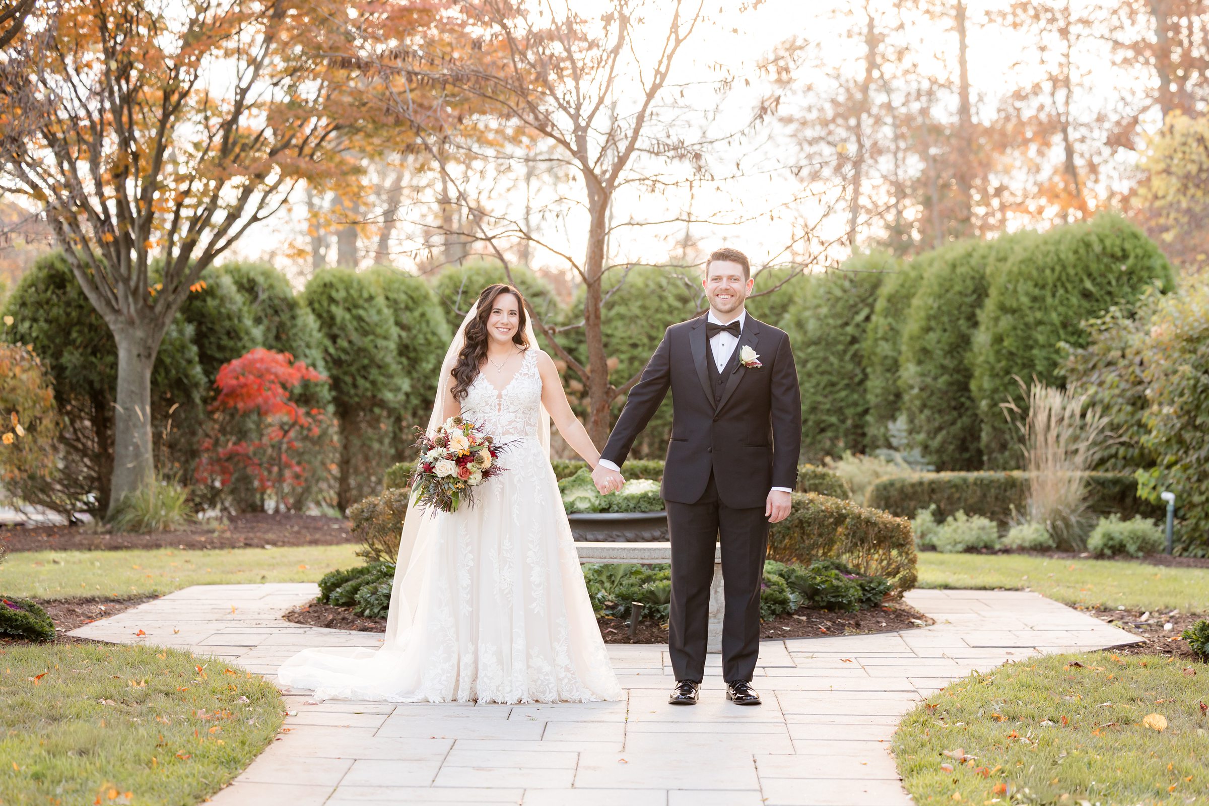 Bride and groom standing together in a symmetrical garden, framed by trees with warm autumn leaves