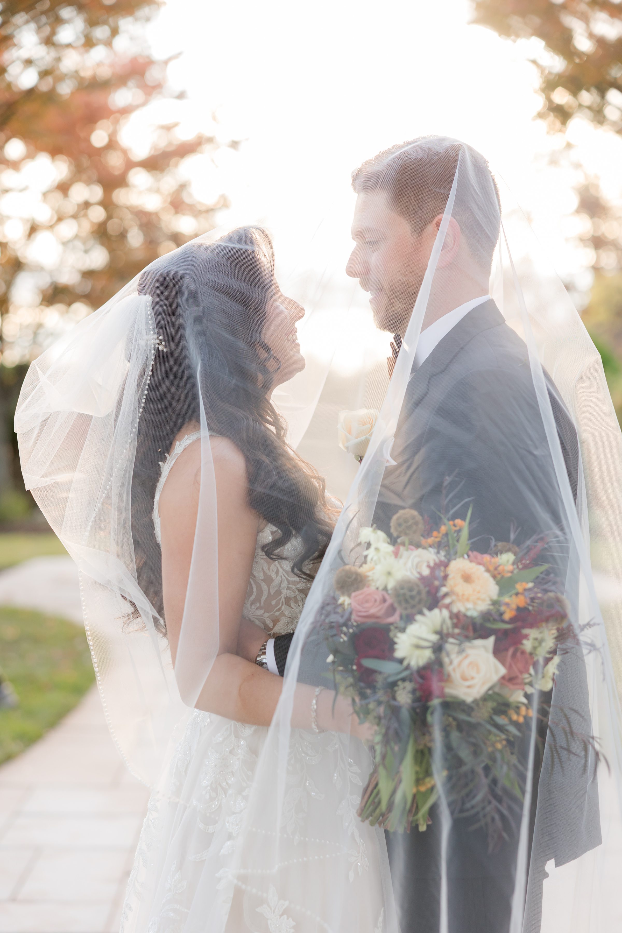 Bride and groom smiling at each other through a sheer veil, surrounded by soft golden light and autumn colors