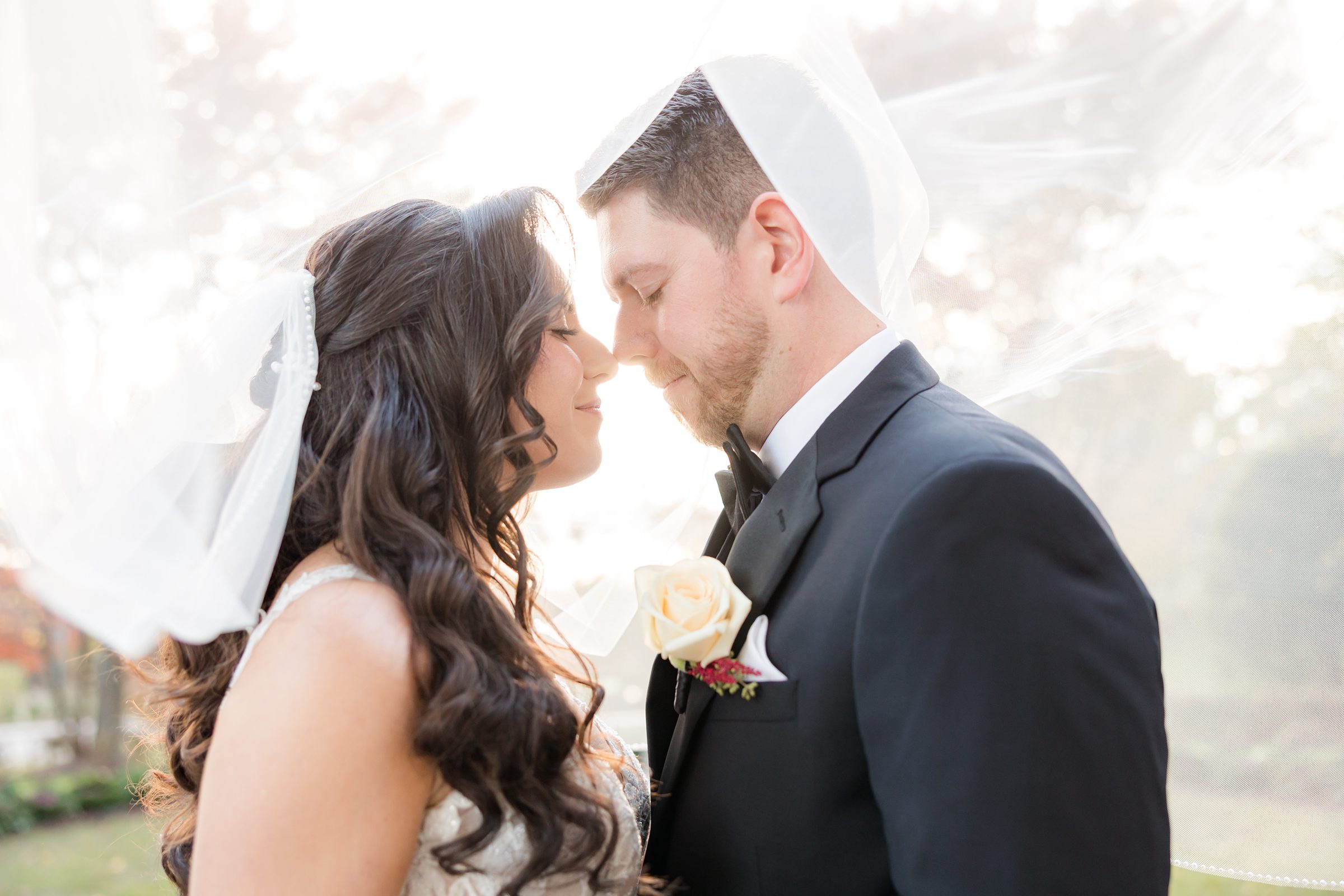 Bride and groom touching foreheads beneath a softly glowing veil, sharing a quiet, romantic moment in warm light