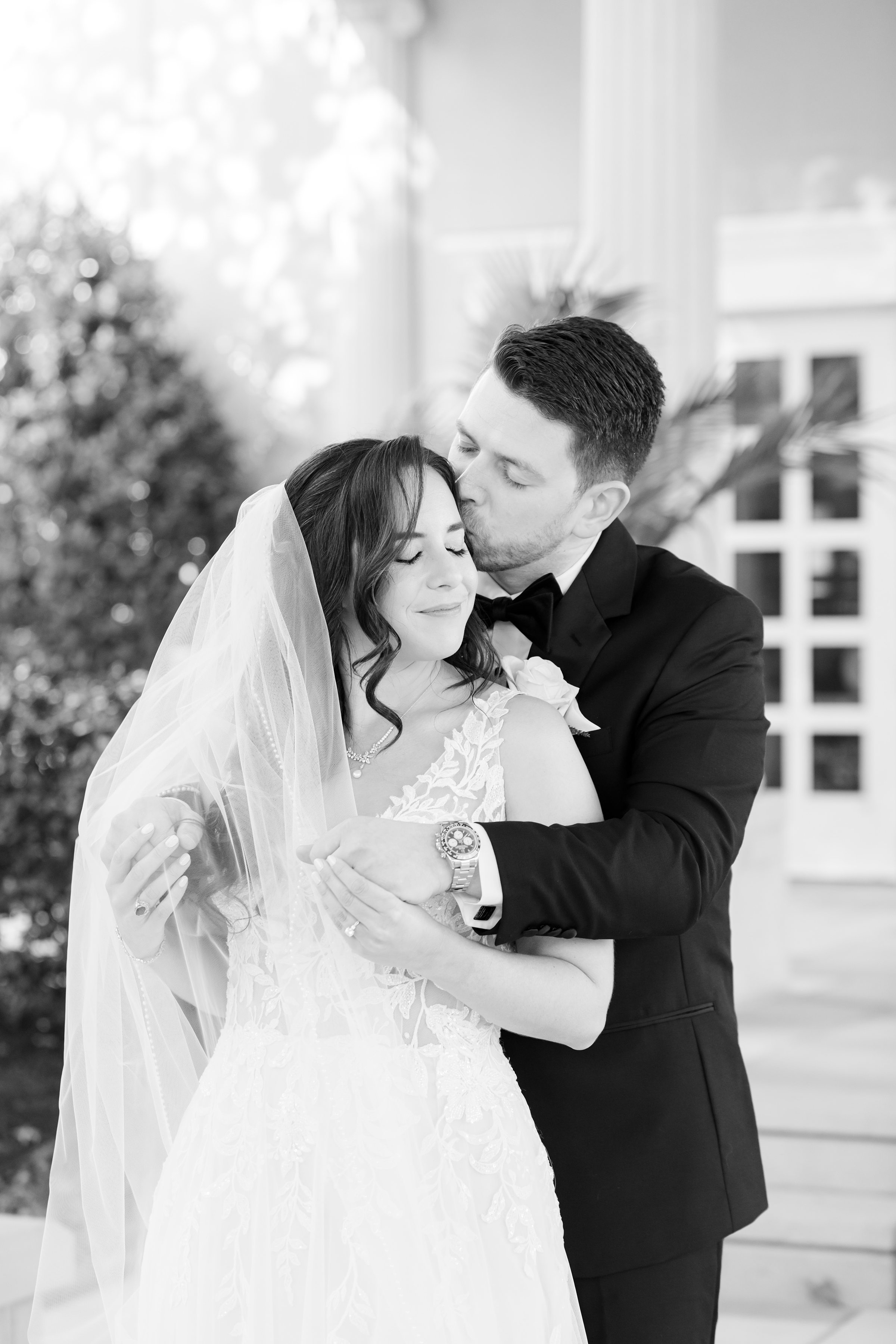Groom wrapping his arms around his bride and kissing her temple as she smiles softly beneath her veil.