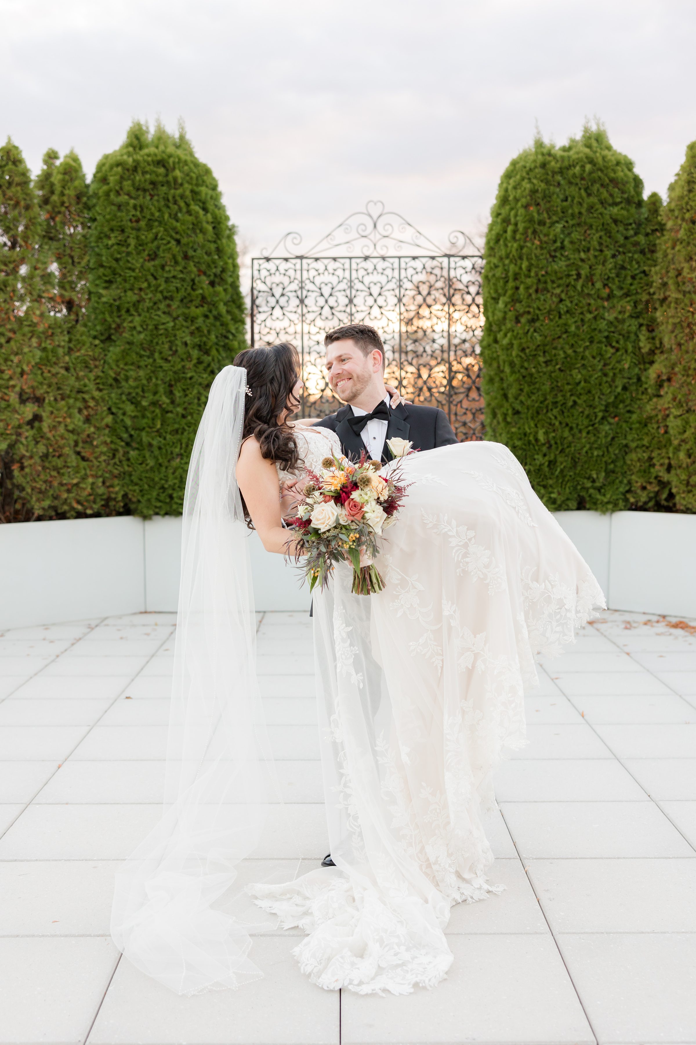 Groom carries his bride in his arms as they smile at each other, surrounded by soft evening light and autumn colors.