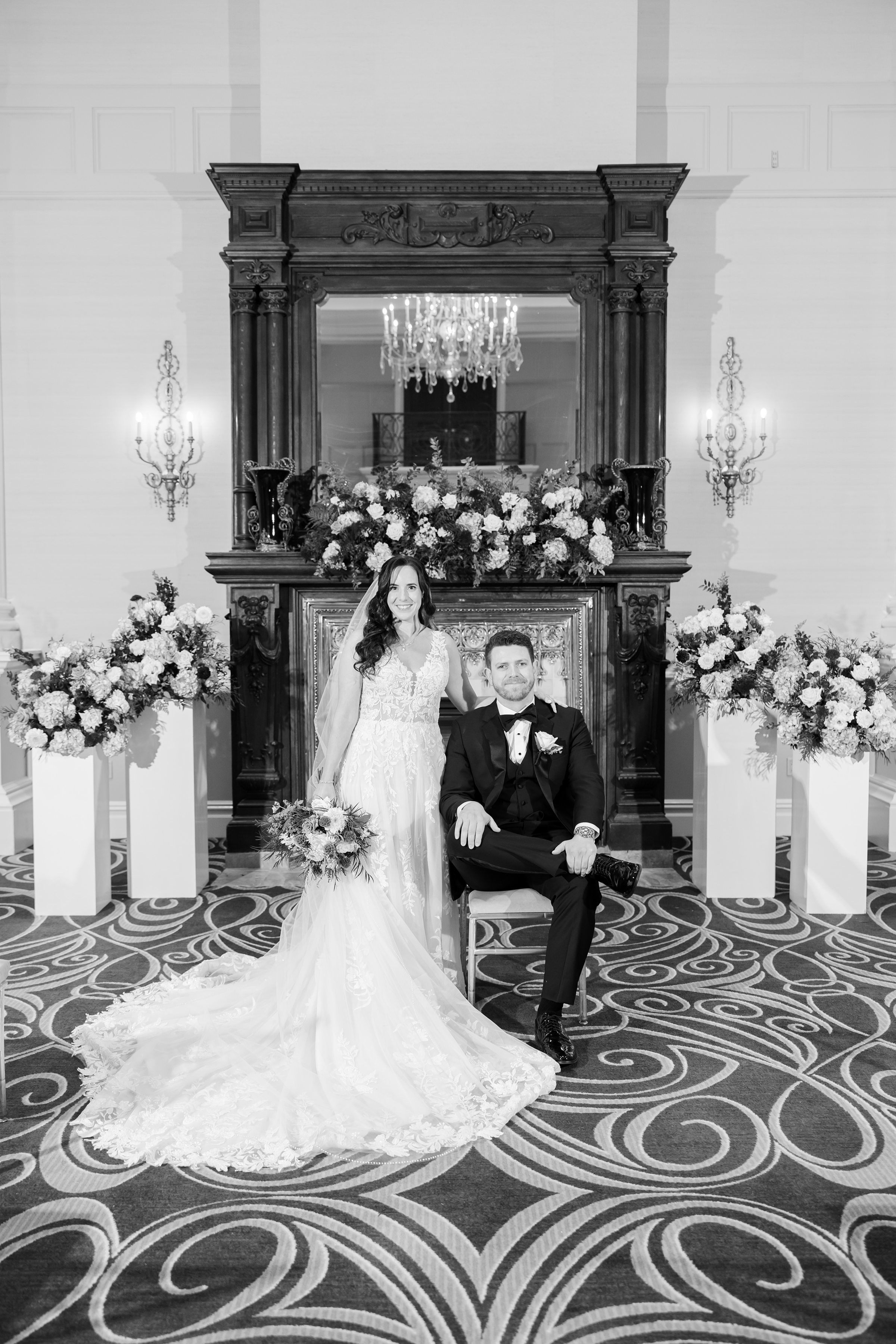 Bride stands beside her seated groom, both composed and radiant in timeless black and white