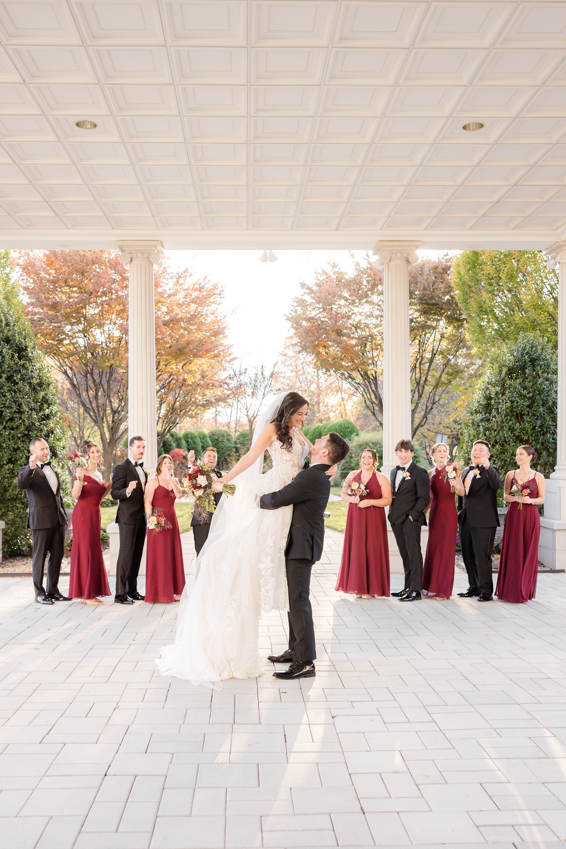 Outdoors beneath a grand structure, the groom lifts his bride high as their wedding party looks on, celebrating their love