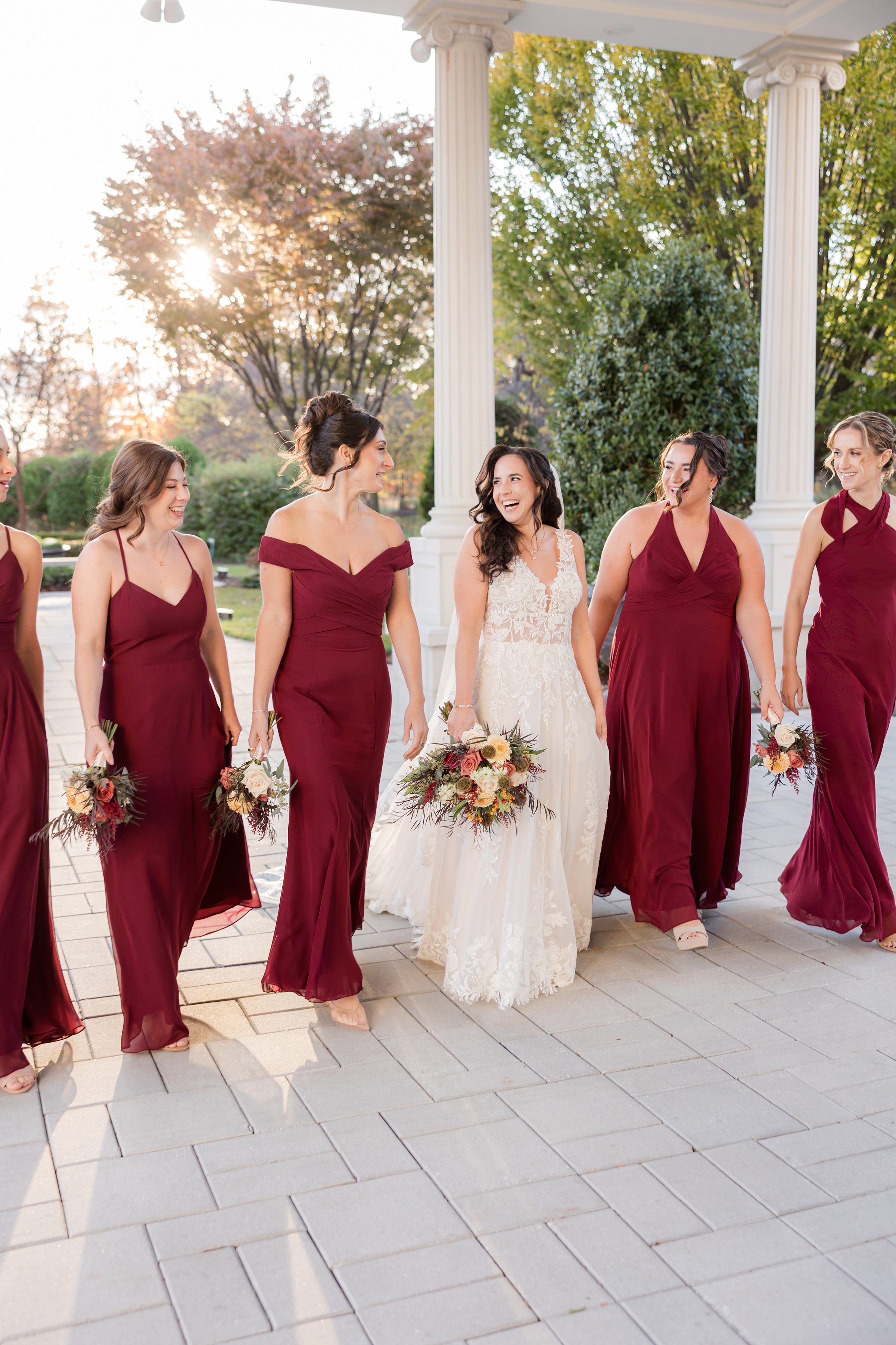 Bride walks smiling with her bridesmaids in burgundy dresses, holding bouquets under warm sunset light.