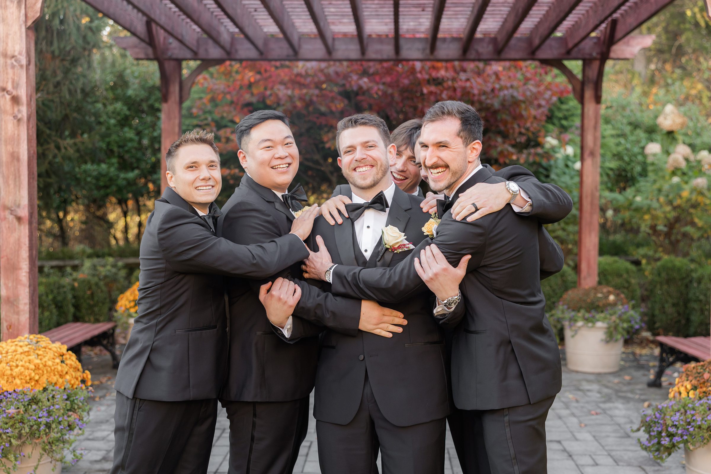 Groom shares a joyful group hug with his groomsmen in black suits beneath a garden pergola
